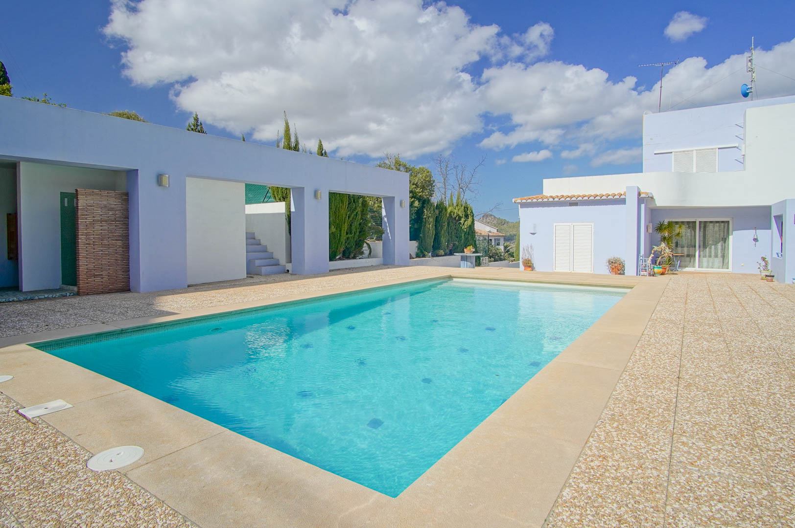 Piscina rectangular rodeada de una amplia terraza de grava y piedra, con muros en tonos lavanda y acceso directo a la vivienda bajo un cielo despejado.
