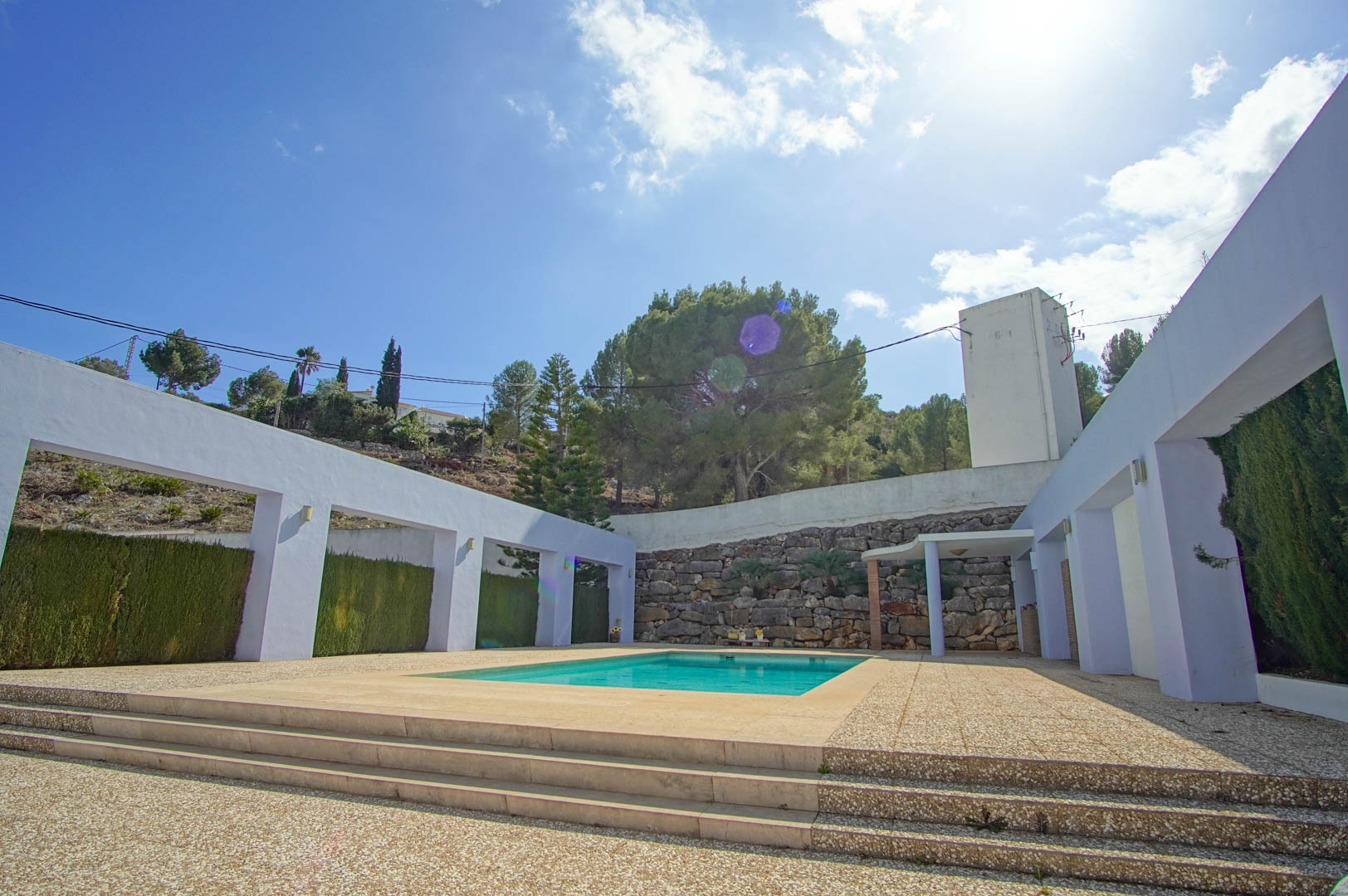 Piscina rectangular rodeada de una amplia terraza de piedra y muros blancos con arcos, enmarcada por un muro de piedra natural y vegetación mediterránea.