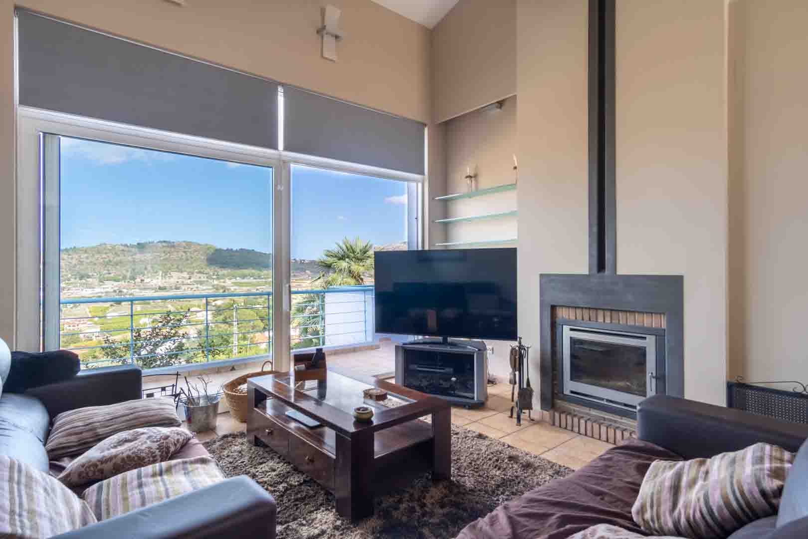 Salón luminoso con chimenea integrada, suelos de gres y grandes ventanales que ofrecen vistas panorámicas al valle y las montañas de Pedreguer.