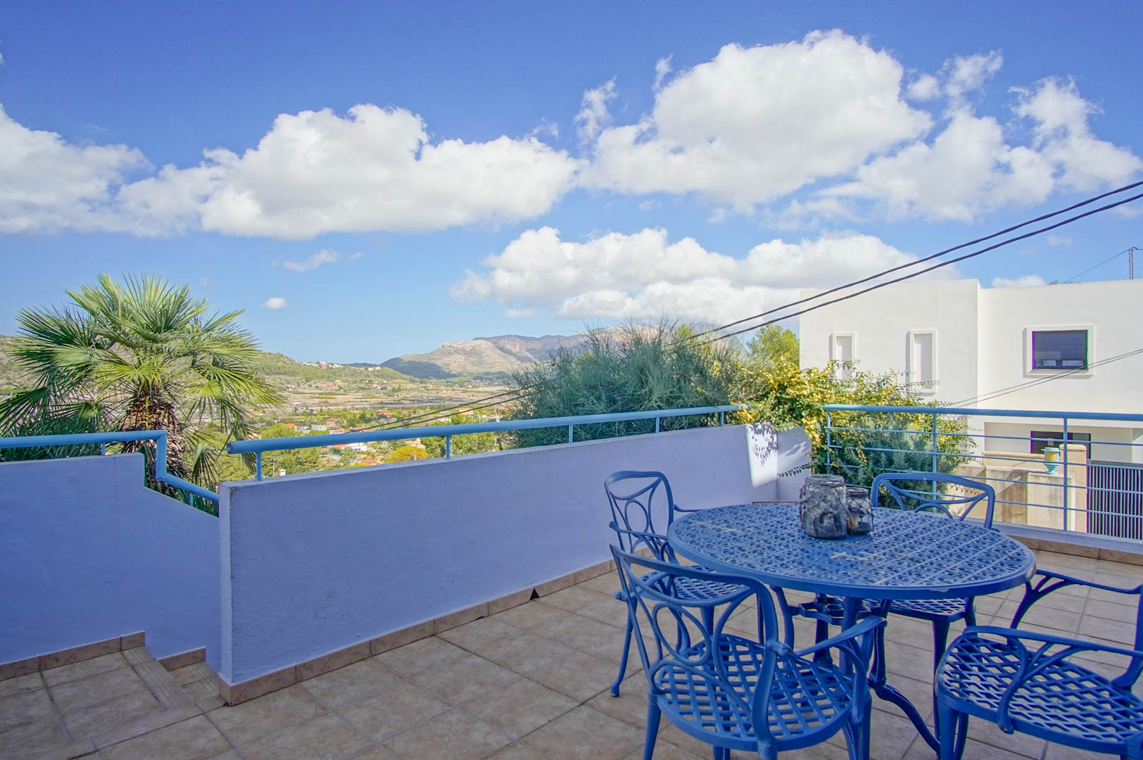 Terraza elevada con mobiliario de forja azul, suelo de gres y vistas panorámicas al valle y las montañas de Pedreguer bajo un cielo despejado.