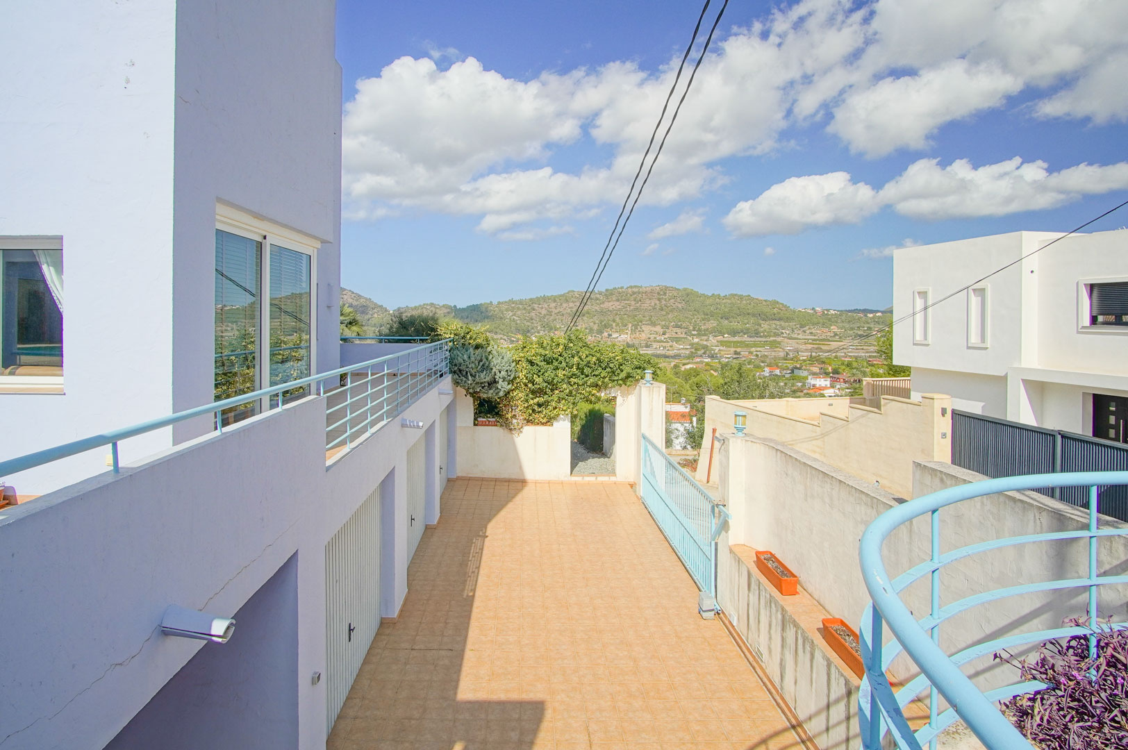 Vista exterior de la zona de acceso con tres garajes privados, suelos cerámicos y barandillas azules, con vistas despejadas a las montañas de Pedreguer.