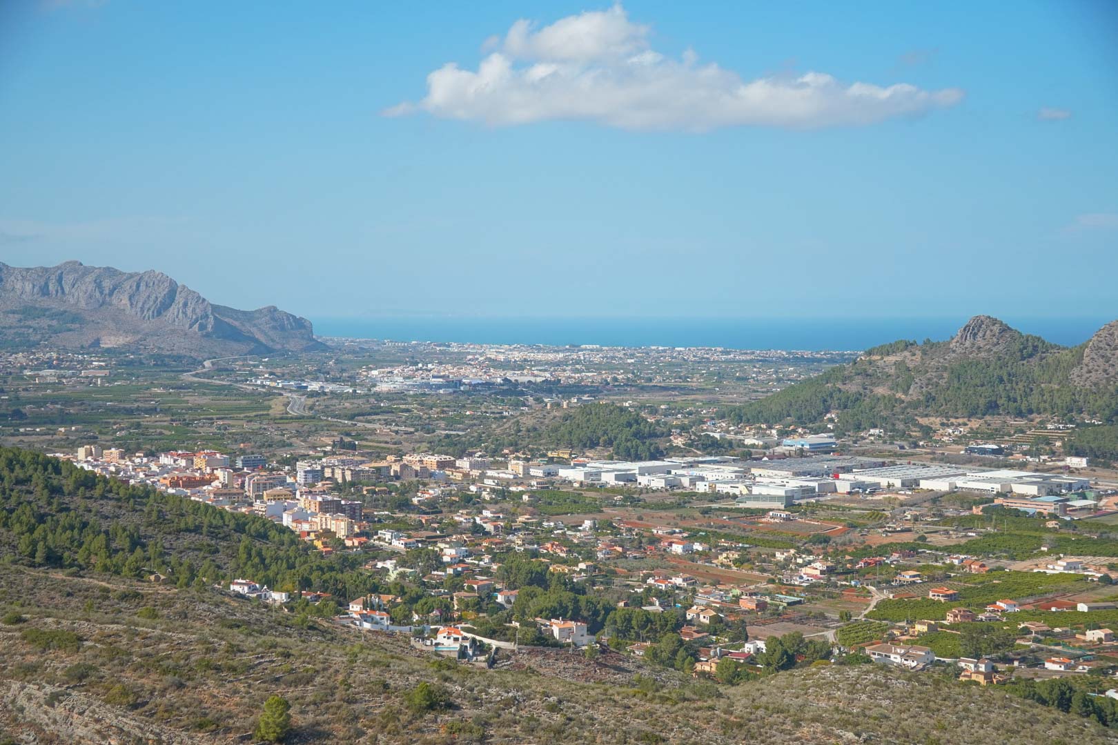 Vista panorámica elevada que muestra el valle de Pedreguer, las formaciones montañosas y el mar Mediterráneo en el horizonte bajo un cielo azul claro.