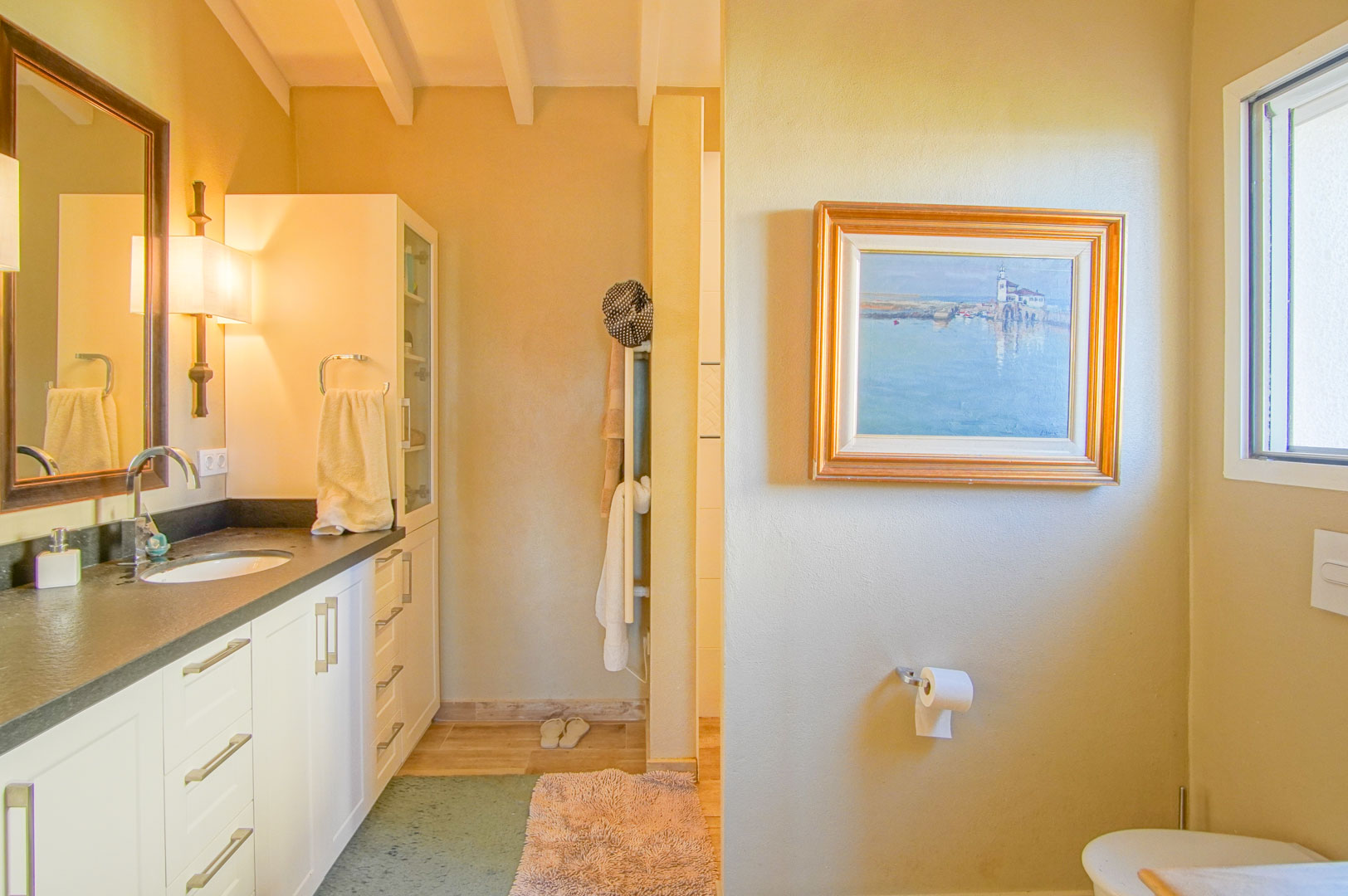 Baño con encimera oscura, lavabo integrado y vigas de madera a la vista. Incluye mobiliario blanco, iluminación cálida y una ventana que aporta luz natural al espacio.