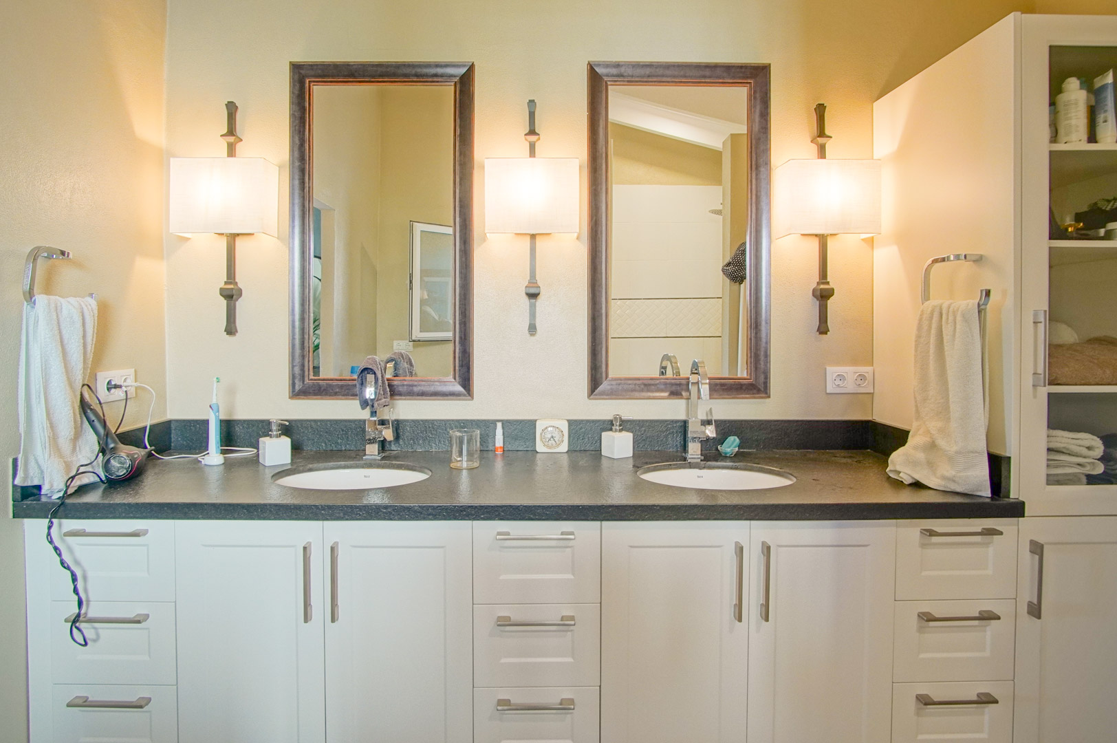 Baño con lavabo doble, encimera de piedra oscura y muebles blancos. Incluye espejos enmarcados y apliques de pared de estilo clásico para una iluminación cálida.