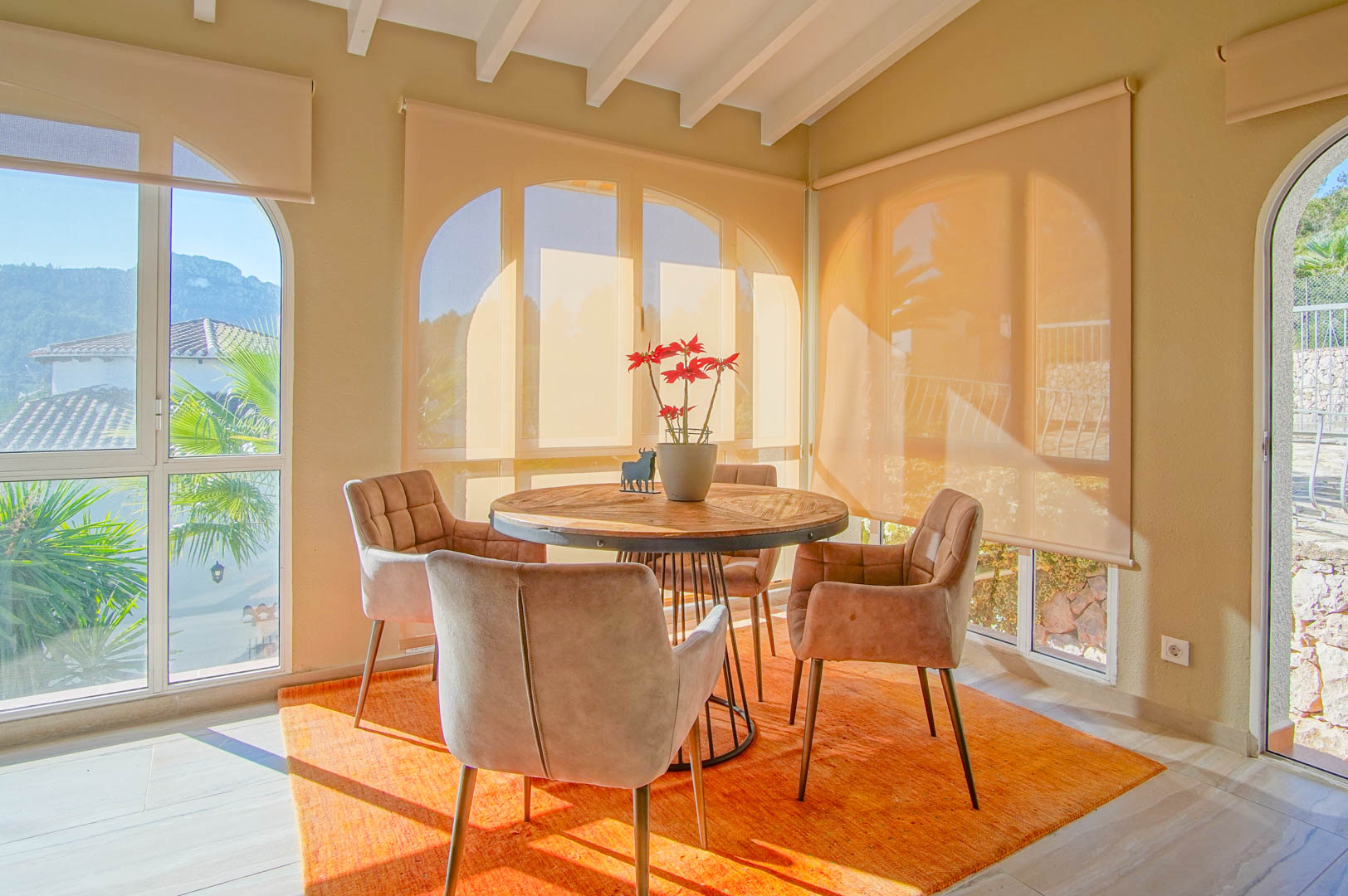 Comedor luminoso con mesa redonda de madera, vigas blancas expuestas y amplios ventanales con vistas a la montaña y persianas enrollables.
