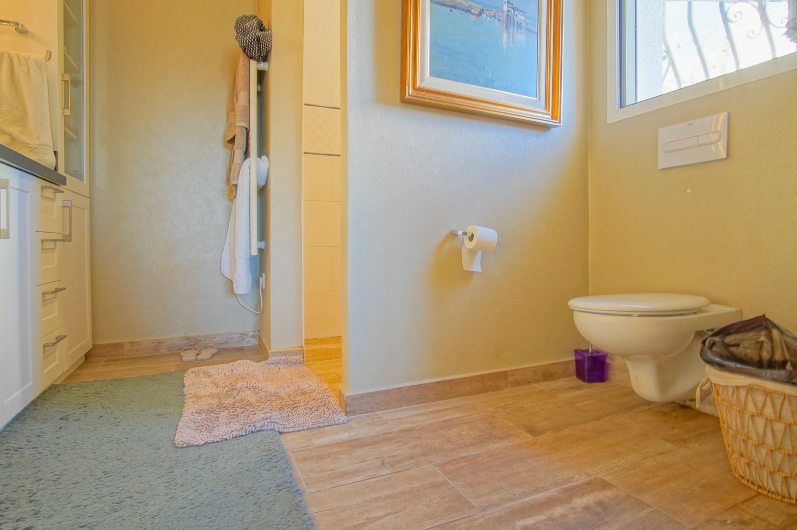 Cuarto de baño con inodoro suspendido, suelos de gres efecto madera y mobiliario blanco. Incluye radiador toallero y acceso a una zona de ducha alicatada.