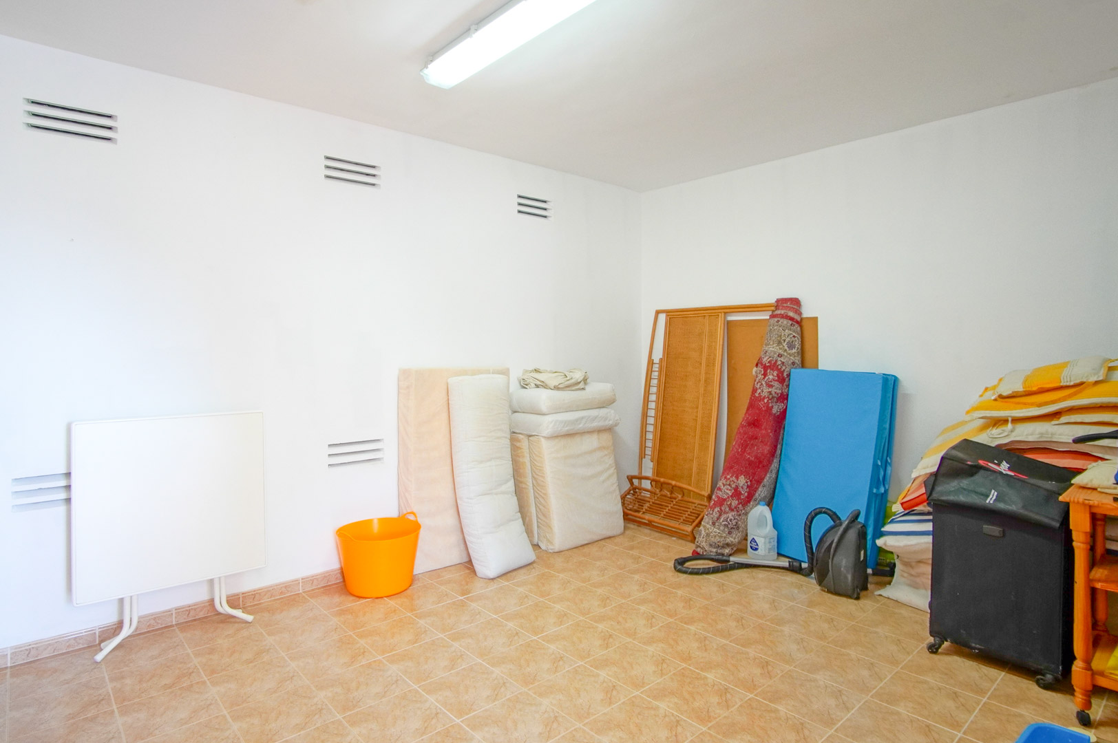 Cuarto trastero funcional con paredes blancas, rejillas de ventilación y suelos cerámicos. Un espacio versátil iluminado por luz fluorescente, ideal para almacenamiento.