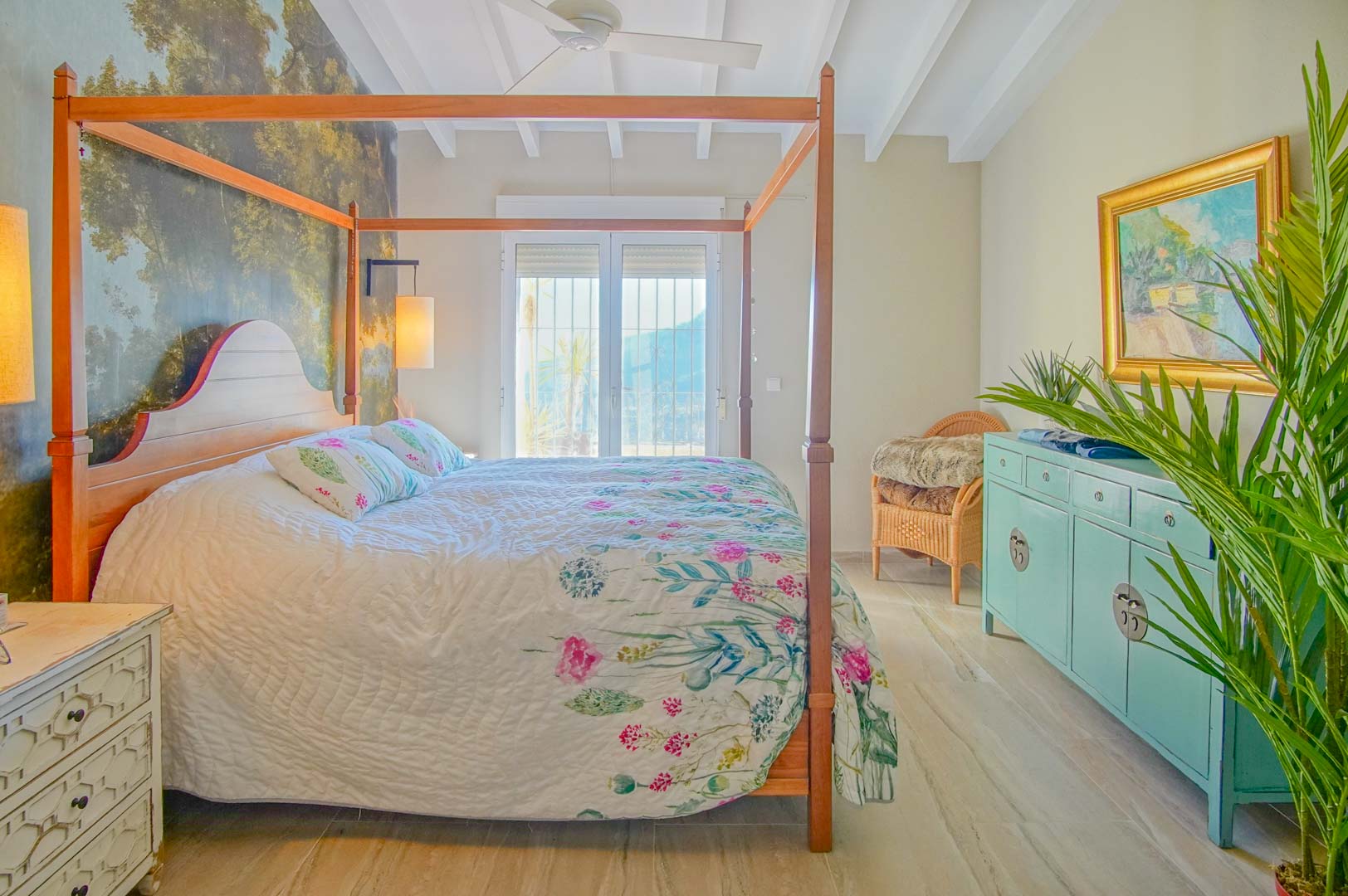 Dormitorio con cama de dosel de madera, mural decorativo de bosque, techo de vigas blancas y acceso exterior a través de puertas acristaladas.