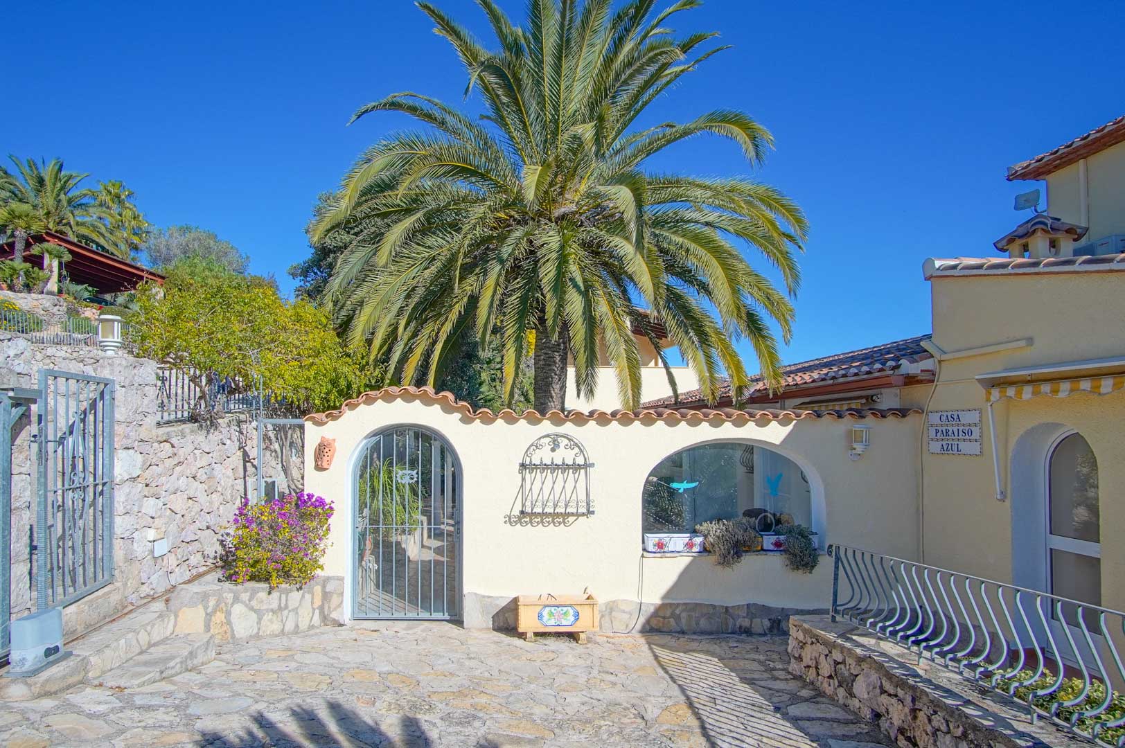 Entrada exterior con pavimento de piedra natural, muros blancos rematados con teja cerámica y una gran palmera central que preside el acceso.