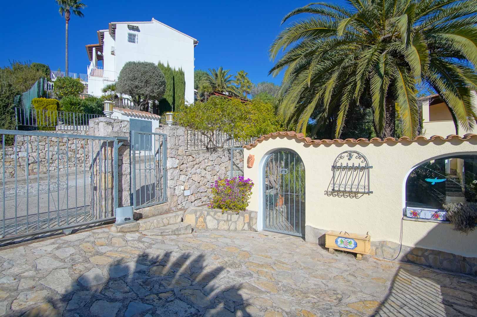 Entrada principal con portón metálico corredero, muros de piedra natural y acceso peatonal arqueado, rodeado de vegetación mediterránea y palmeras.