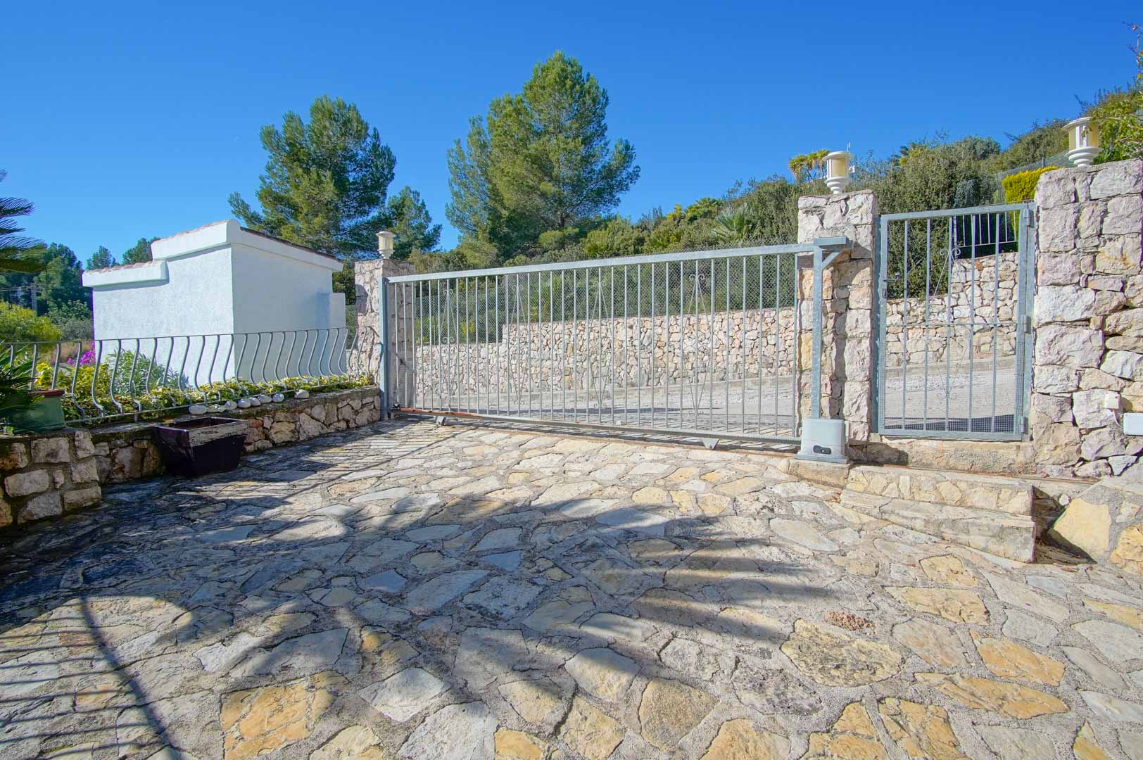 Entrada principal con puerta corredera automática y acceso peatonal, rodeada de muros de piedra natural y suelo de losas irregulares en un entorno arbolado.