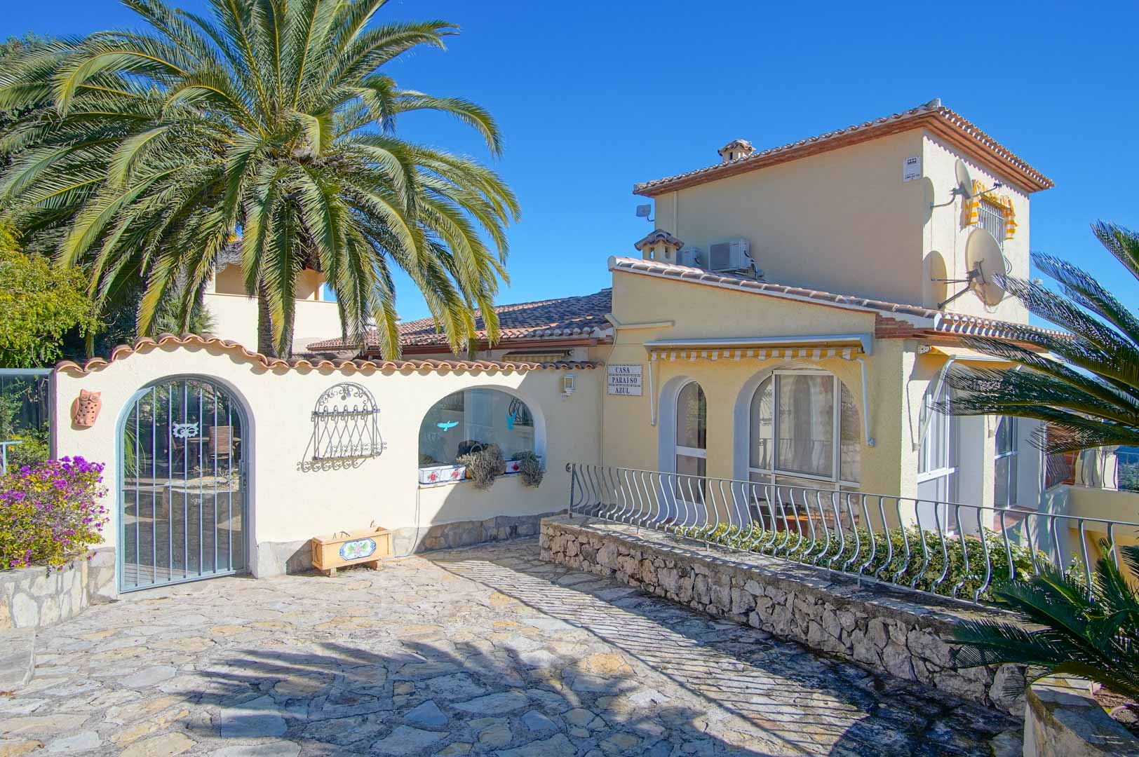 Fachada principal de la villa con entrada empedrada, muros de piedra natural, arcos decorativos y vegetación mediterránea bajo un cielo despejado.