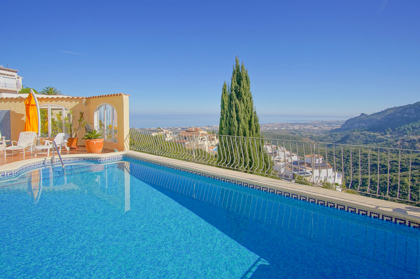 Piscina exterior con borde de mosaico azul, rodeada por una terraza con tumbonas y vistas panorámicas hacia el mar Mediterráneo y las montañas de Monte Pego.