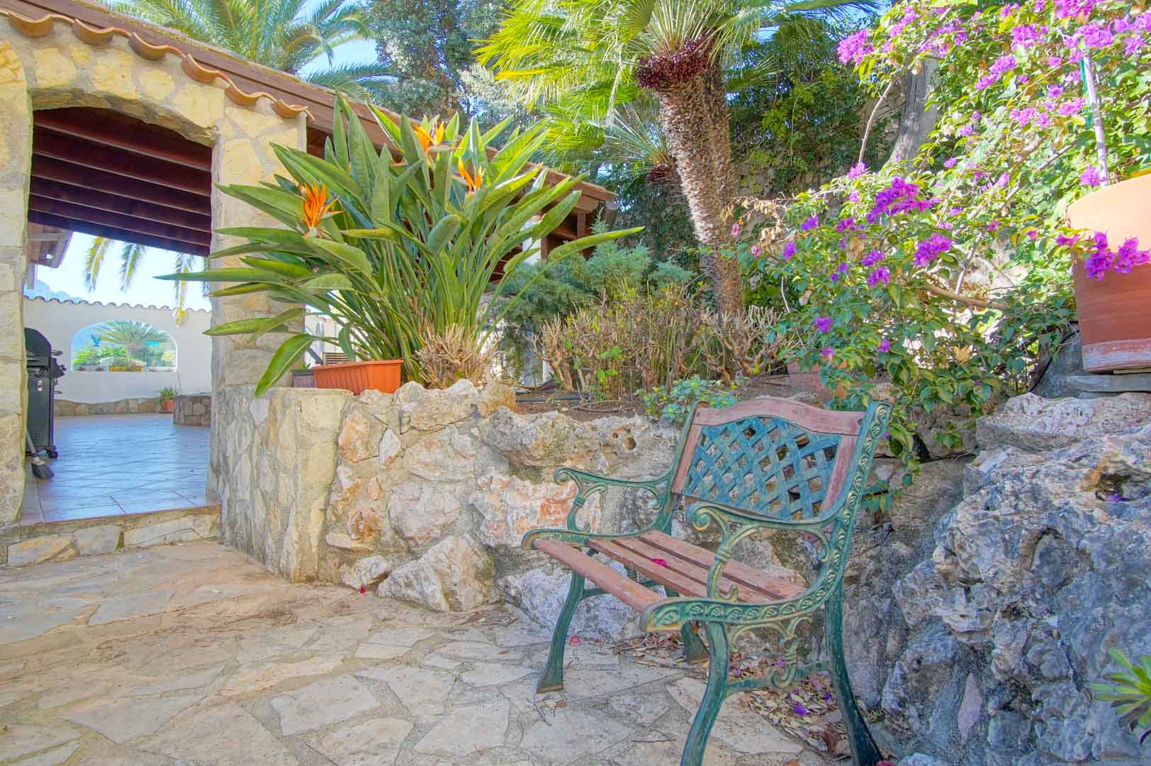 Rincón de descanso con banco de forja y madera, muros de piedra natural y vegetación mediterránea que precede a una terraza cubierta con vigas de madera.