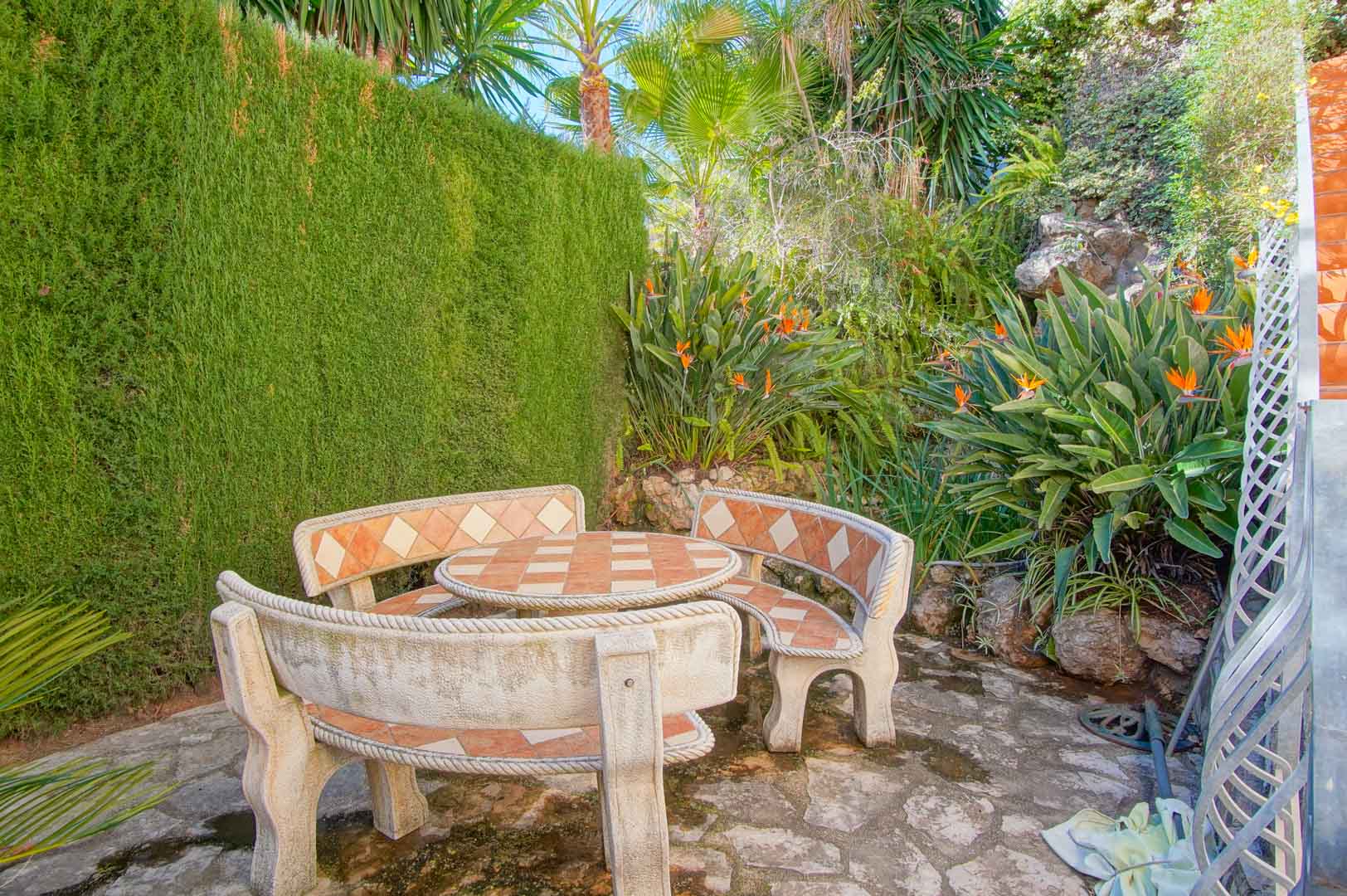 Rincón exterior con mesa y bancos de piedra decorados con azulejos, rodeado de vegetación mediterránea, palmeras y aves del paraíso en un entorno privado.