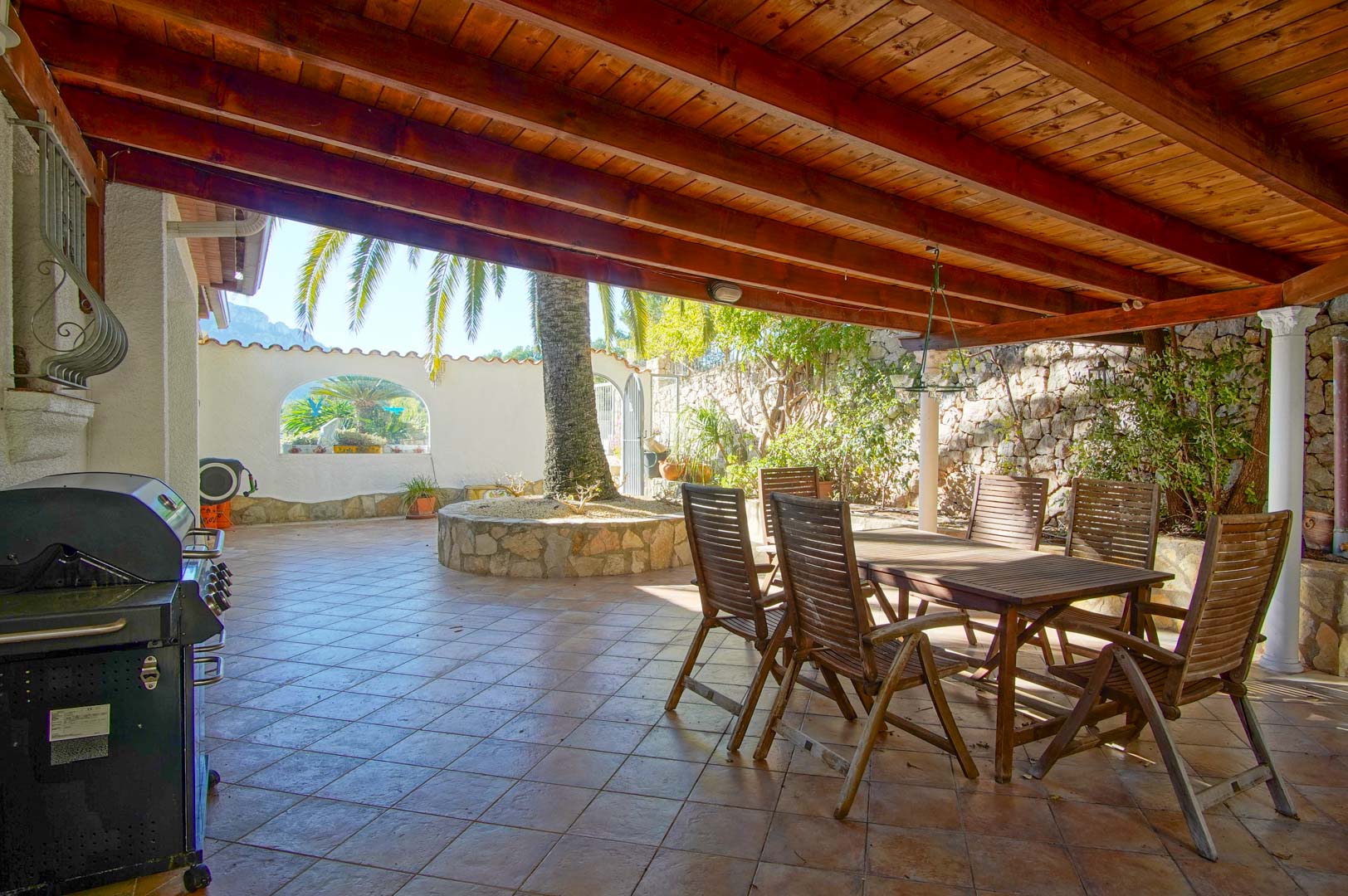 Terraza cubierta con vigas de madera, mesa de comedor exterior, barbacoa y jardinera circular de piedra con palmera, rodeada de muros de piedra natural.