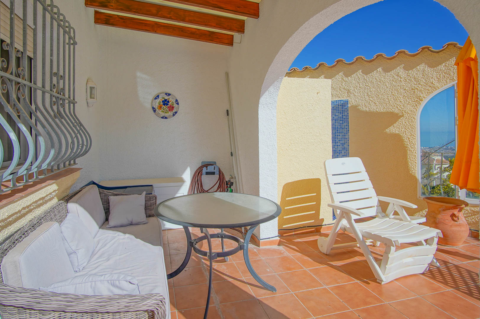 Terraza cubierta con vigas de madera, suelos de terracota y zona de estar que conecta con un solárium con vistas al mar y columna de ducha de mosaico.