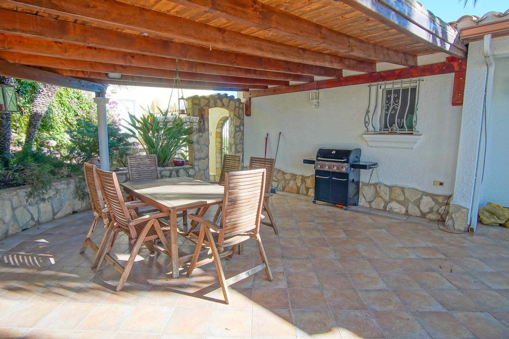 Terraza cubierta con vigas de madera, zona de comedor exterior y barbacoa de gas, sobre suelo cerámico y zócalo de piedra natural.