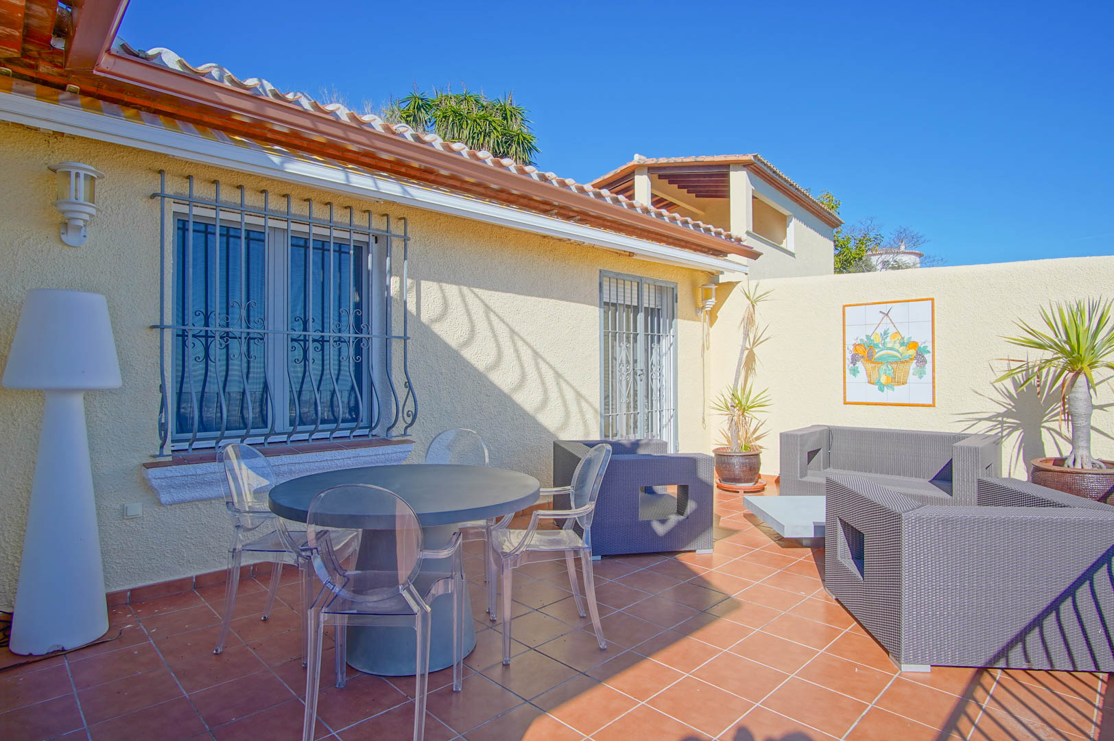 Terraza exterior con suelo de gres, mesa circular con sillas transparentes y zona de estar con sofás de ratán sintético gris bajo un cielo despejado.