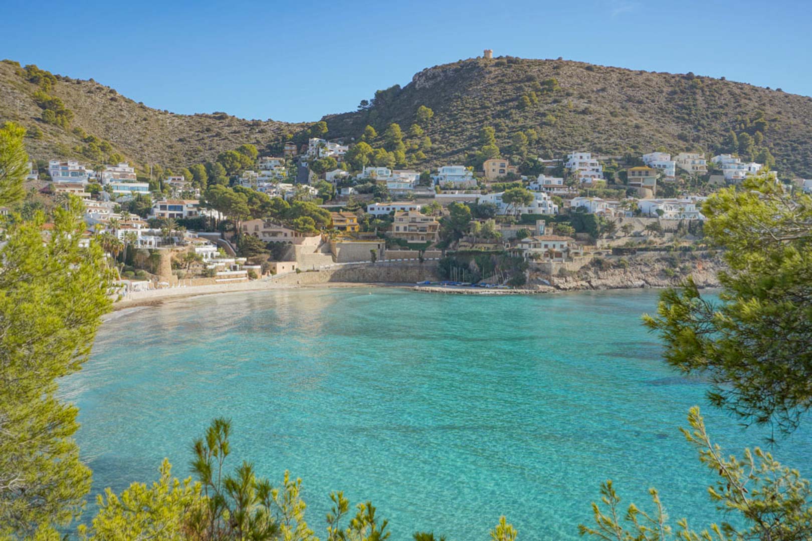 Vista panorámica de la bahía de El Portet en Moraira, con sus aguas cristalinas de color turquesa rodeadas de colinas residenciales y vegetación mediterránea.