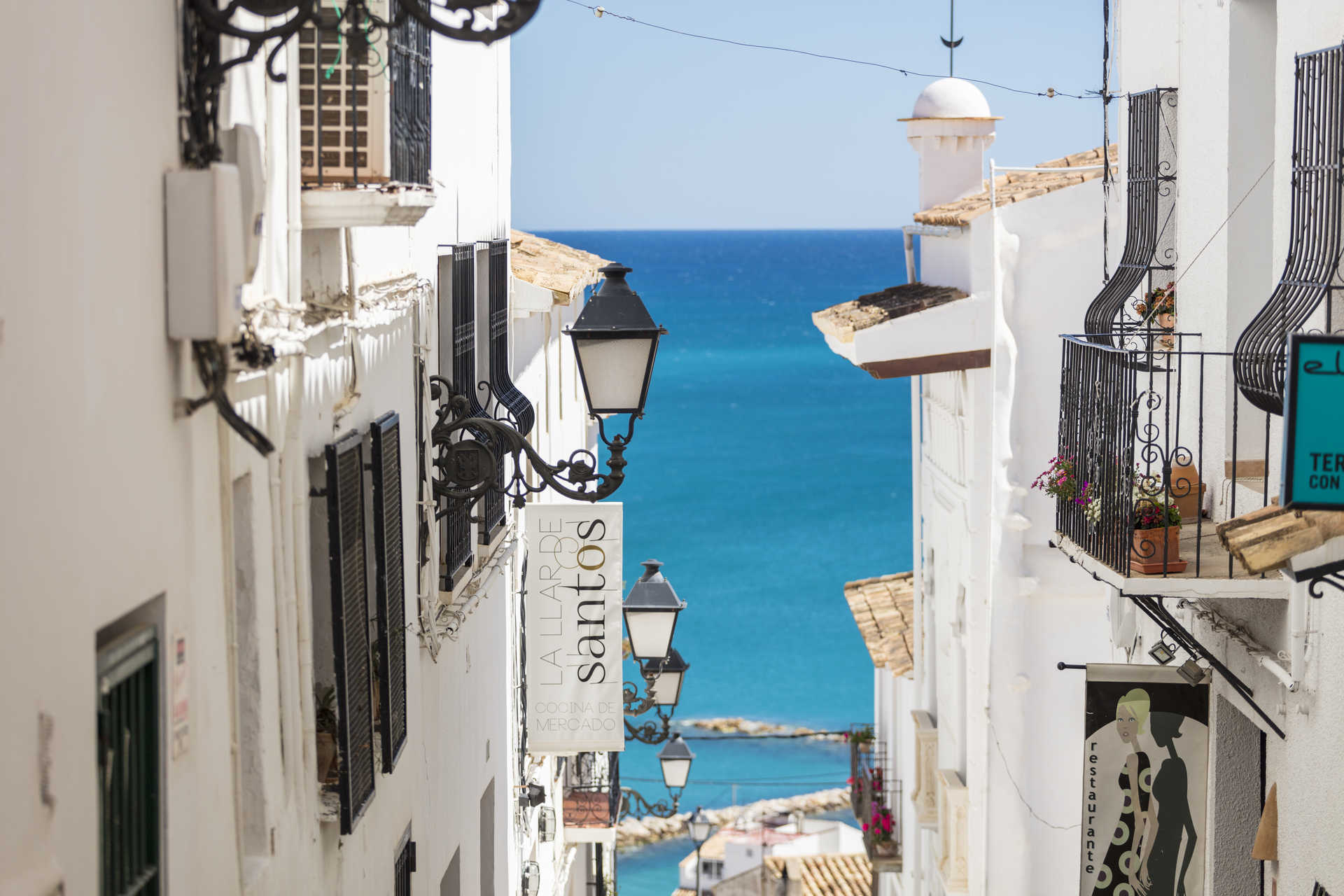 Calle estrecha de fachadas blancas y balcones de forja en el casco antiguo, con vistas al mar Mediterráneo y farolas de estilo tradicional.