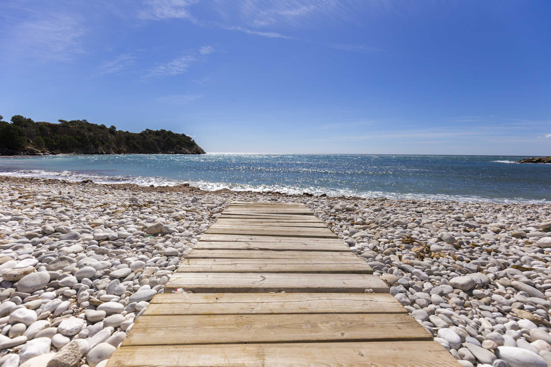 Pasarela de madera sobre playa de cantos blancos con vistas directas al mar Mediterráneo y la costa de Altea bajo un cielo despejado.