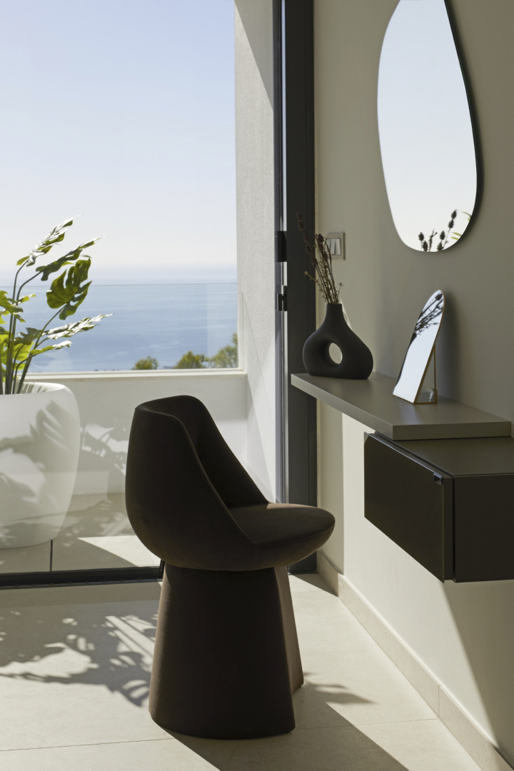 Tocador de diseño con espejo orgánico y silla escultórica, situado frente a un gran ventanal con vistas panorámicas al mar Mediterráneo.