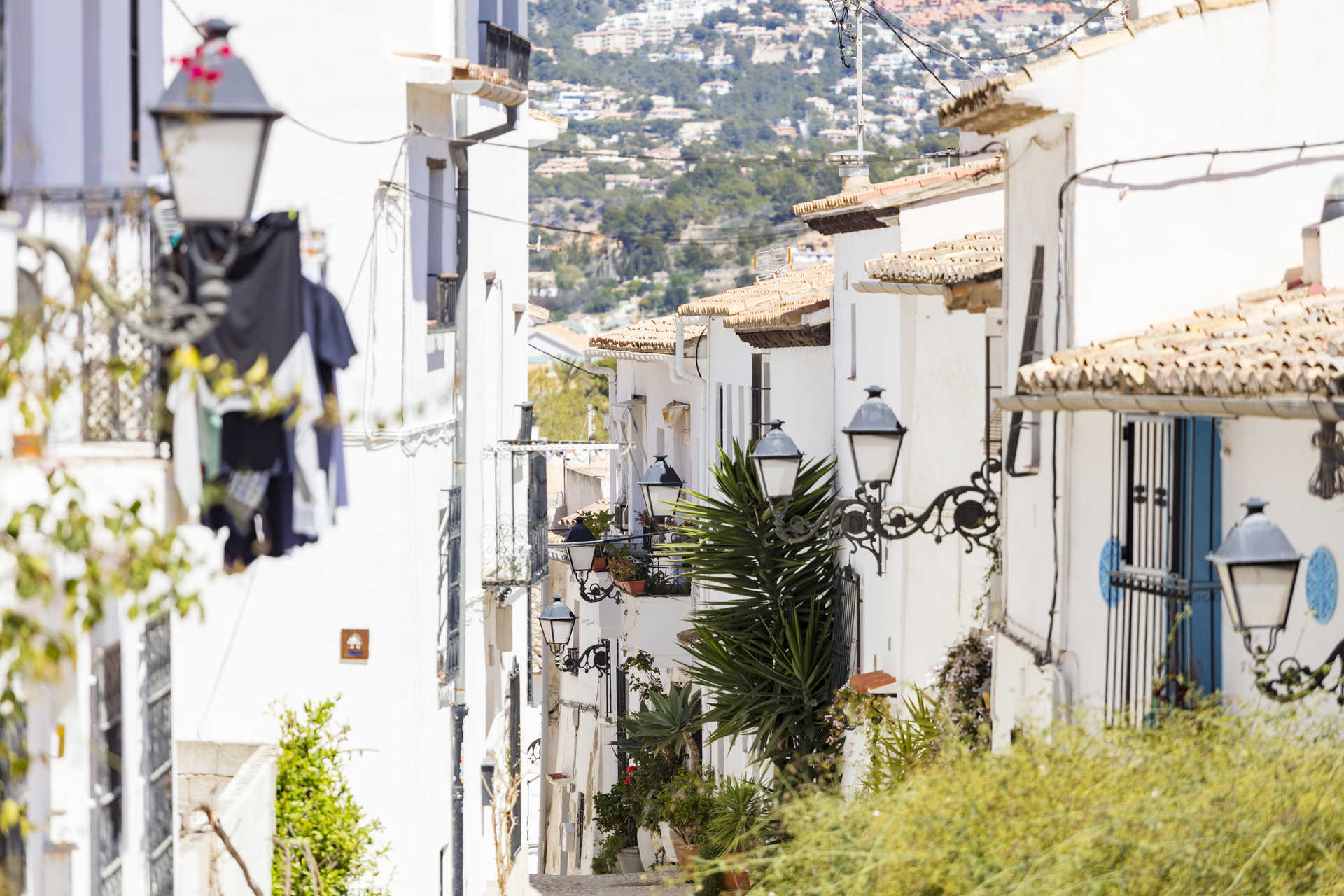 Vista de una calle estrecha tradicional en Altea con fachadas blancas, farolas de hierro forjado y tejados de teja cerámica bajo la luz del sol.