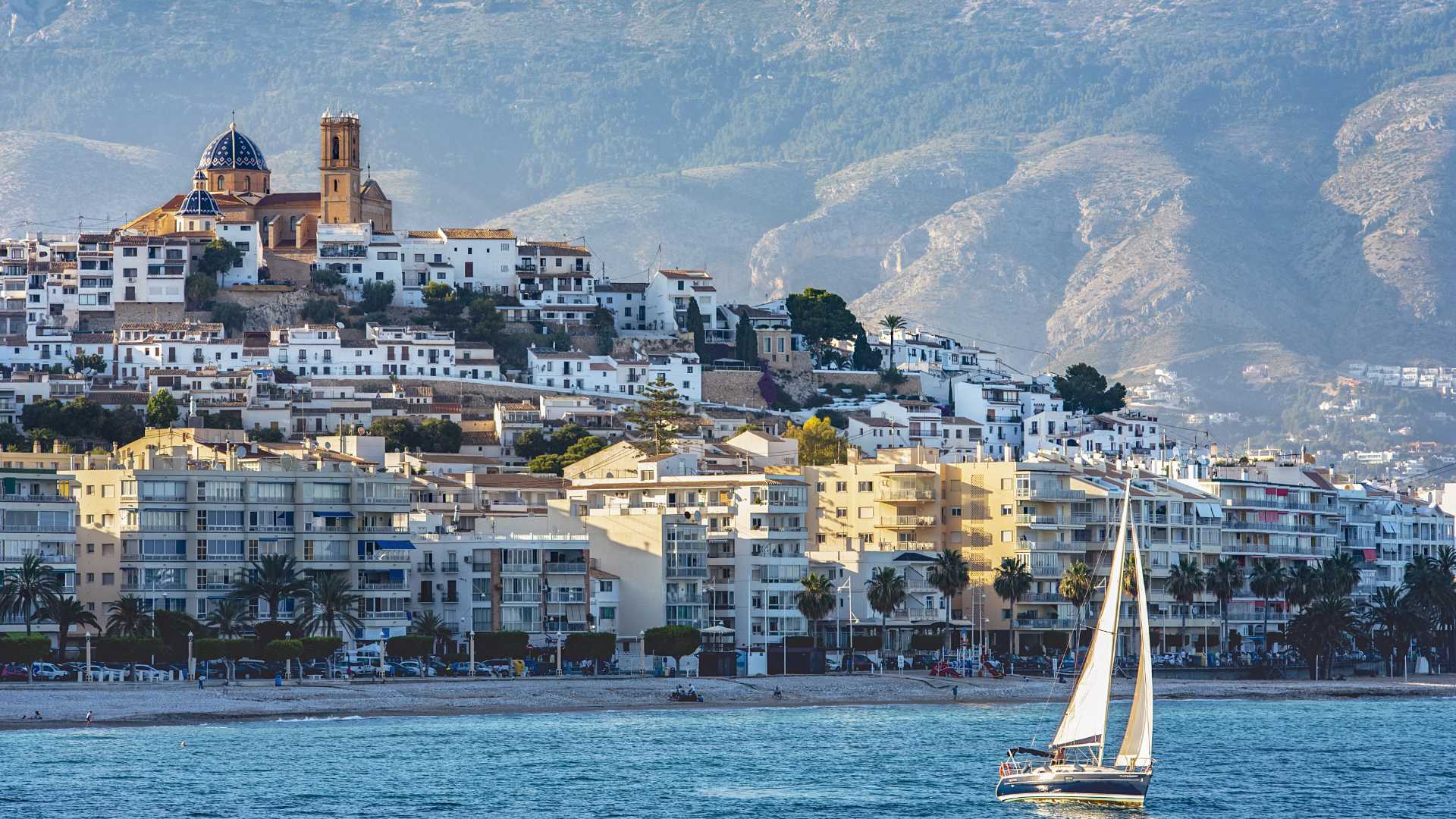 Vista panorámica del casco antiguo de Altea con su icónica iglesia de cúpulas azules, el mar Mediterráneo y un velero navegando frente a la costa.
