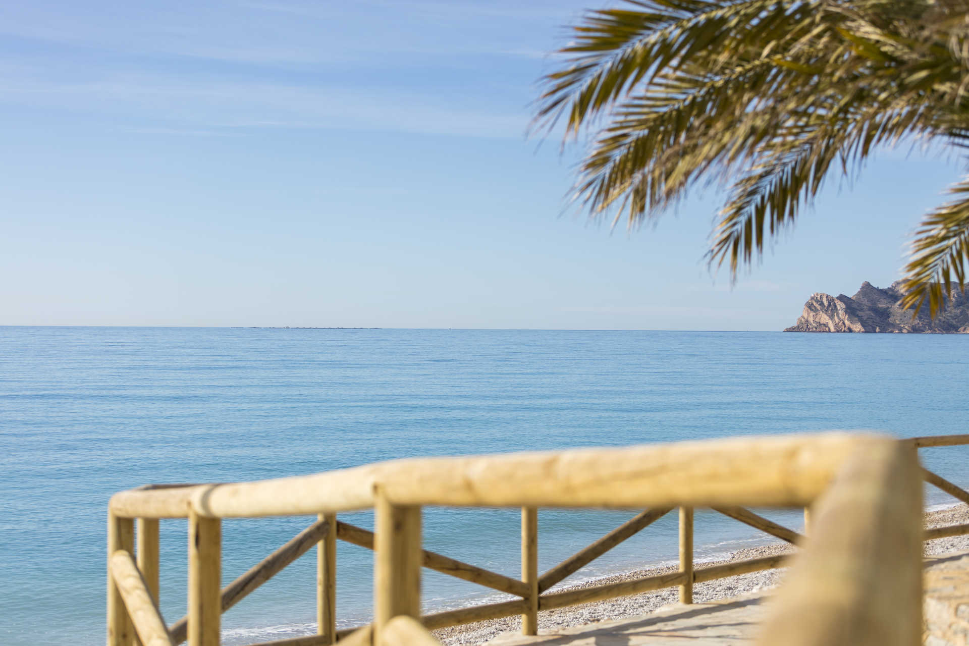 Vistas panorámicas al mar Mediterráneo desde un sendero costero con barandilla de madera, palmeras y el perfil de la costa de Altea al fondo.
