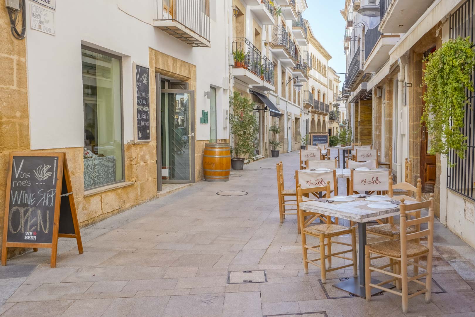 Calle peatonal en el centro histórico de Jávea con fachadas de piedra tosca, balcones de hierro forjado y terrazas de restaurantes con mobiliario de madera.