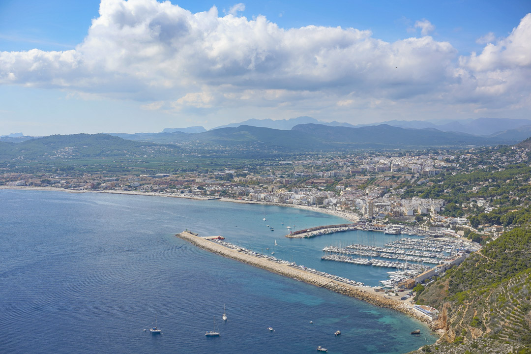 Vista panorámica del puerto de Jávea y su bahía, mostrando el club náutico, la costa mediterránea y las montañas del interior bajo un cielo despejado.