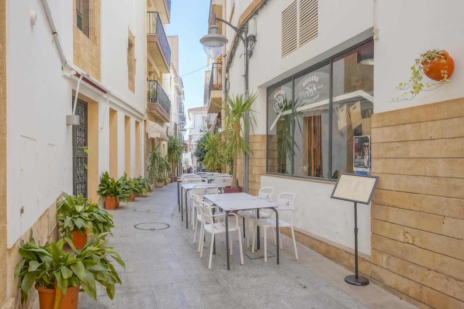 Calle peatonal en el centro histórico de Jávea con fachadas blancas, detalles en piedra tosca y terrazas al aire libre rodeadas de plantas mediterráneas.