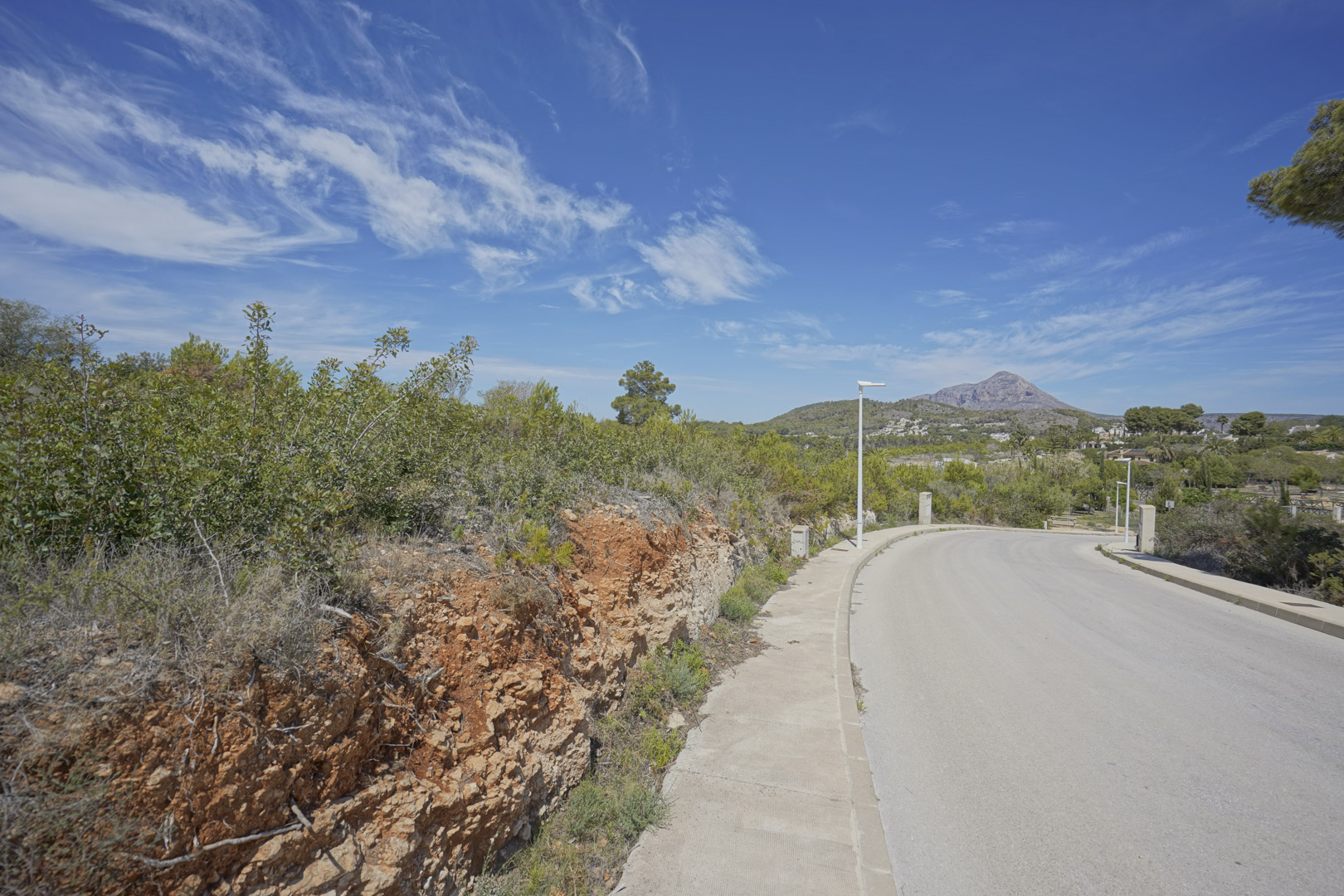 Vista de la calle de acceso en la zona de Valsol, Jávea, con el macizo del Montgó al fondo y vegetación mediterránea autóctona bajo un cielo despejado.