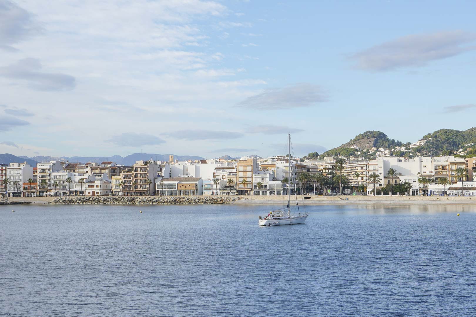 Vista panorámica de la costa de Jávea con un velero fondeado, destacando la arquitectura mediterránea frente al mar y el relieve montañoso al fondo.