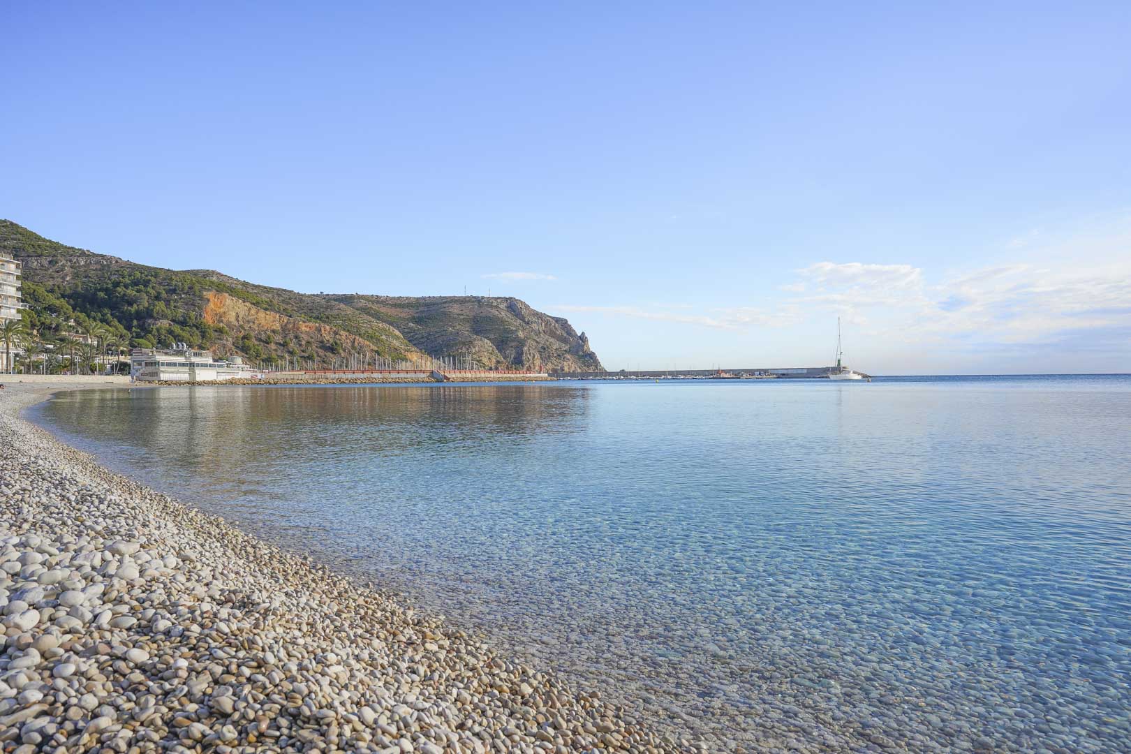 Vista panorámica de la playa de guijarros en Jávea, con aguas cristalinas y el imponente Cabo de San Antonio al fondo bajo un cielo despejado.