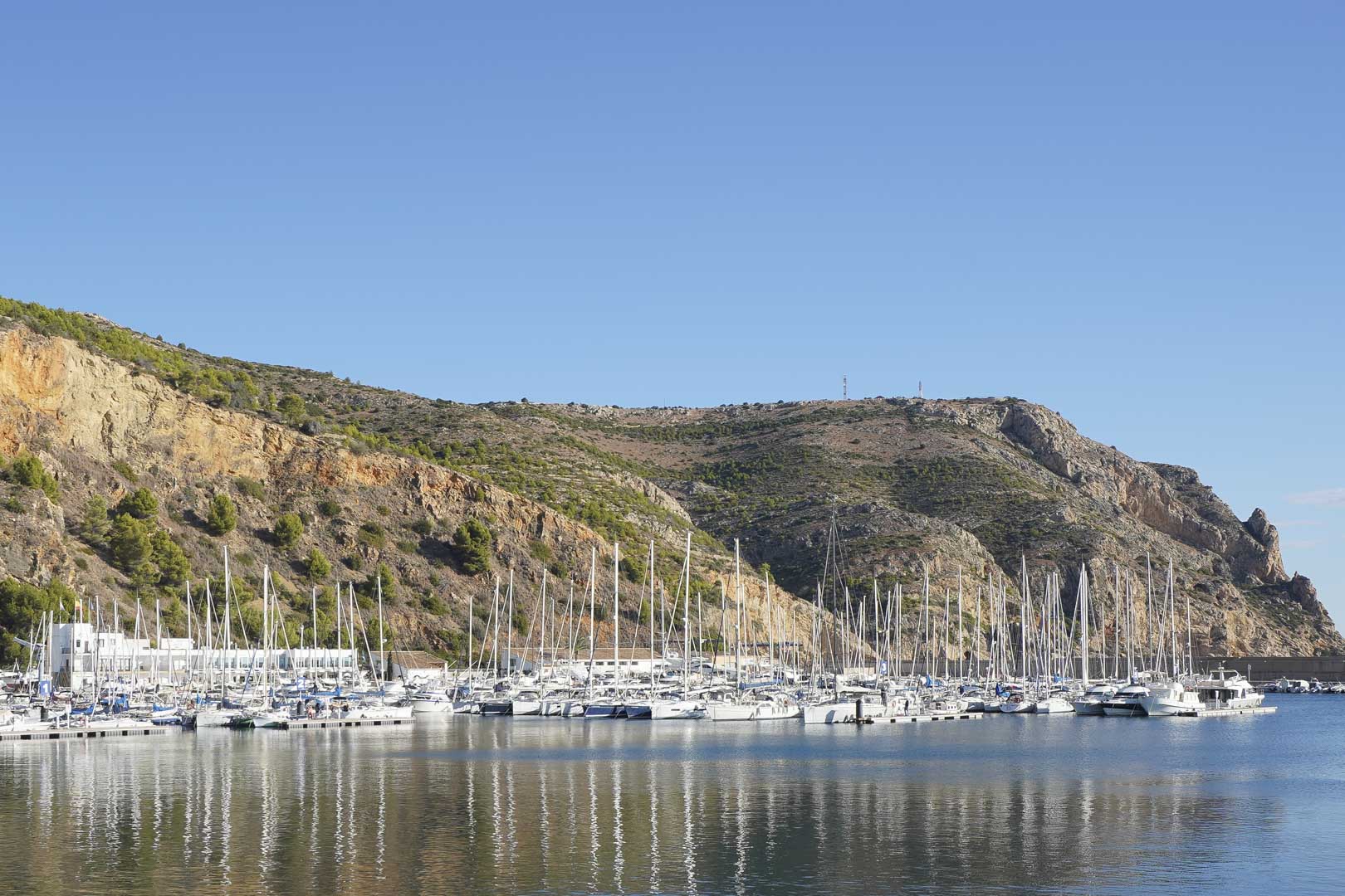 Vista panorámica del puerto deportivo de Jávea, con numerosos veleros atracados frente a los acantilados mediterráneos bajo un cielo despejado.