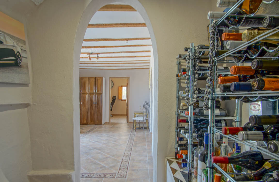 Bodega con estanterías metálicas que conecta mediante un arco con una estancia de vigas de madera vistas, suelos cerámicos decorados y armarios de madera.