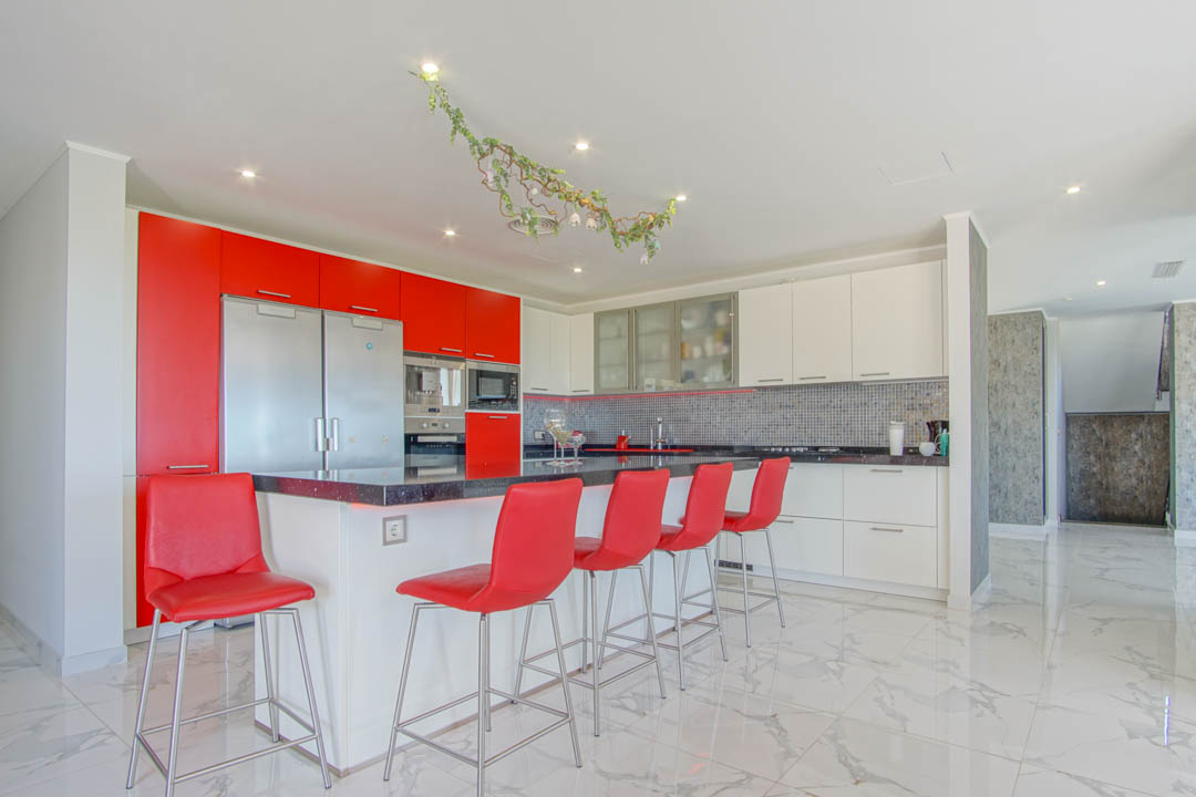Cocina moderna con isla de granito negro, taburetes rojos y armarios de alto brillo. Presenta suelos de mármol blanco y electrodomésticos de acero inoxidable integrados.
