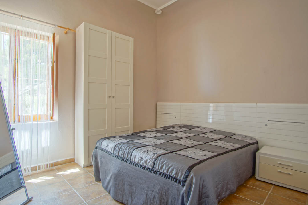 Dormitorio con suelos de baldosa cerámica, armario blanco independiente y cabecero con mesitas integradas bajo luz natural suave.