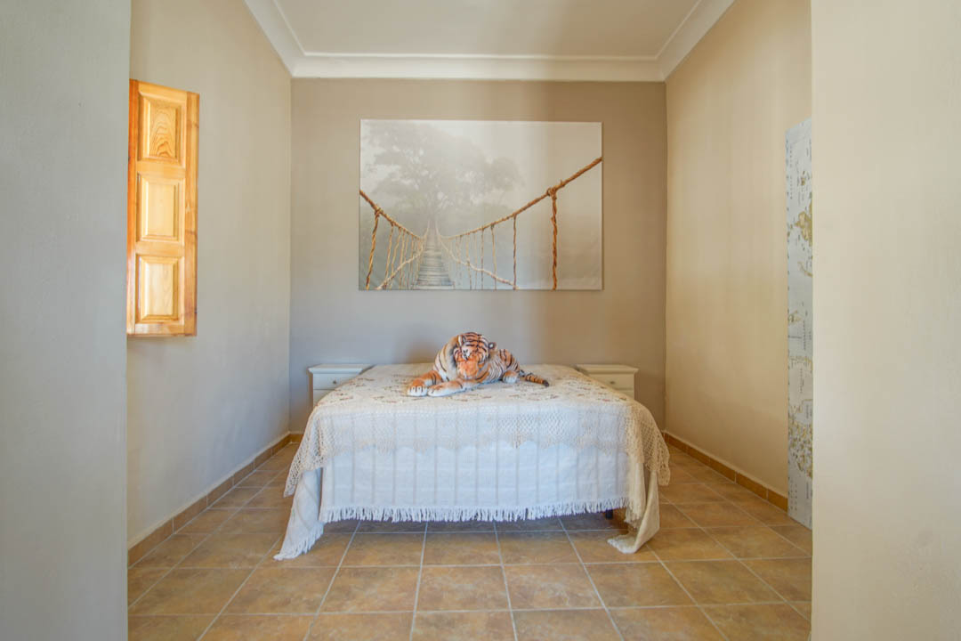 Dormitorio con suelos de gres cerámico, cama doble flanqueada por mesitas blancas y una ventana con contraventanas de madera natural.