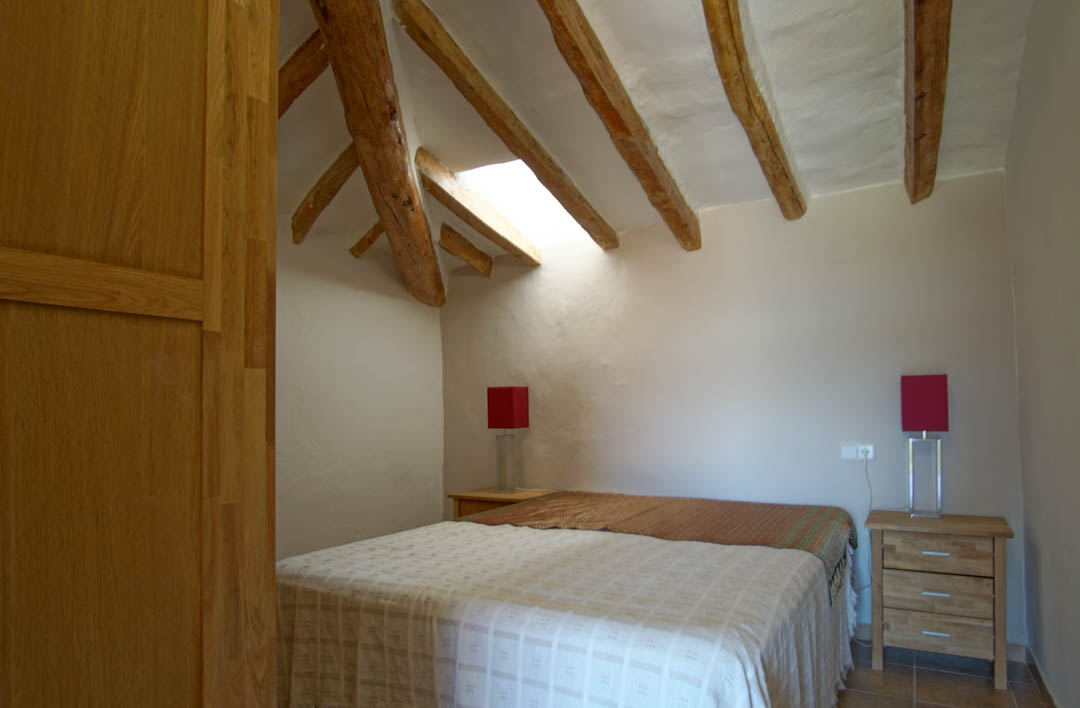 Dormitorio rústico con vigas de madera a la vista, claraboya que aporta luz natural y paredes de yeso blanco. Incluye mesitas de noche de madera y lámparas de acento rojas.