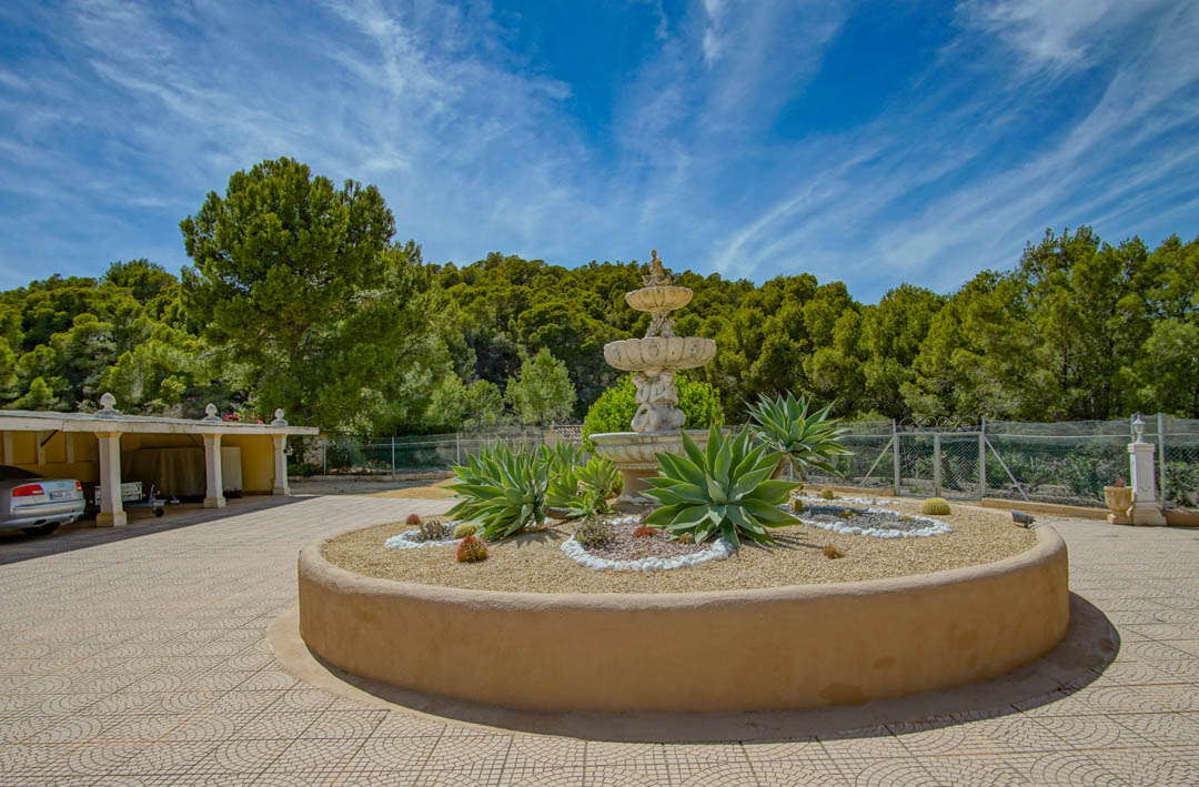 Entrada principal con fuente clásica de tres niveles y parterre circular con agaves, junto a un amplio aparcamiento techado y entorno forestal.