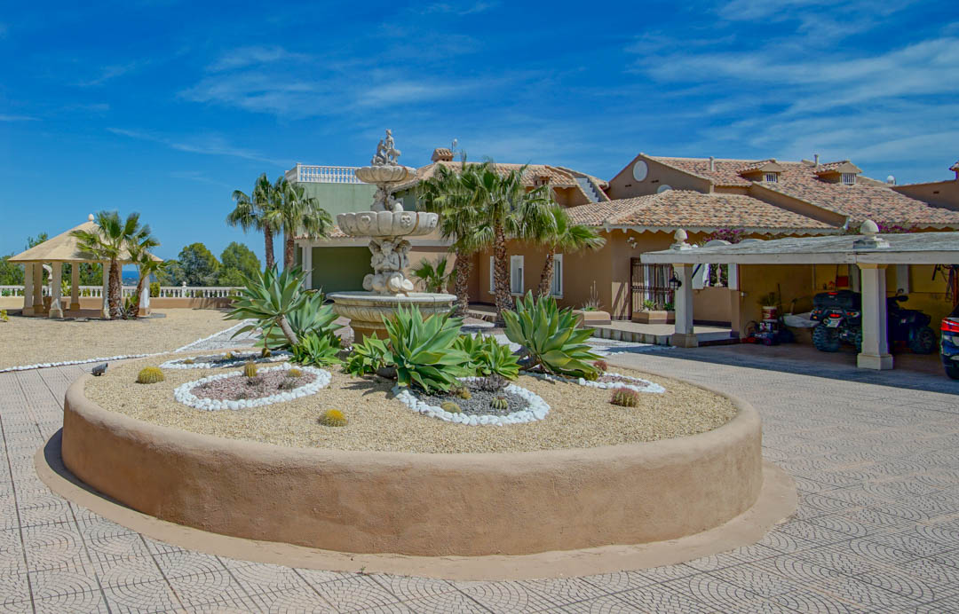 Entrada principal con fuente ornamental de piedra, parterre circular con agaves y cactus, zona de aparcamiento cubierto y pabellón de jardín bajo un cielo despejado.