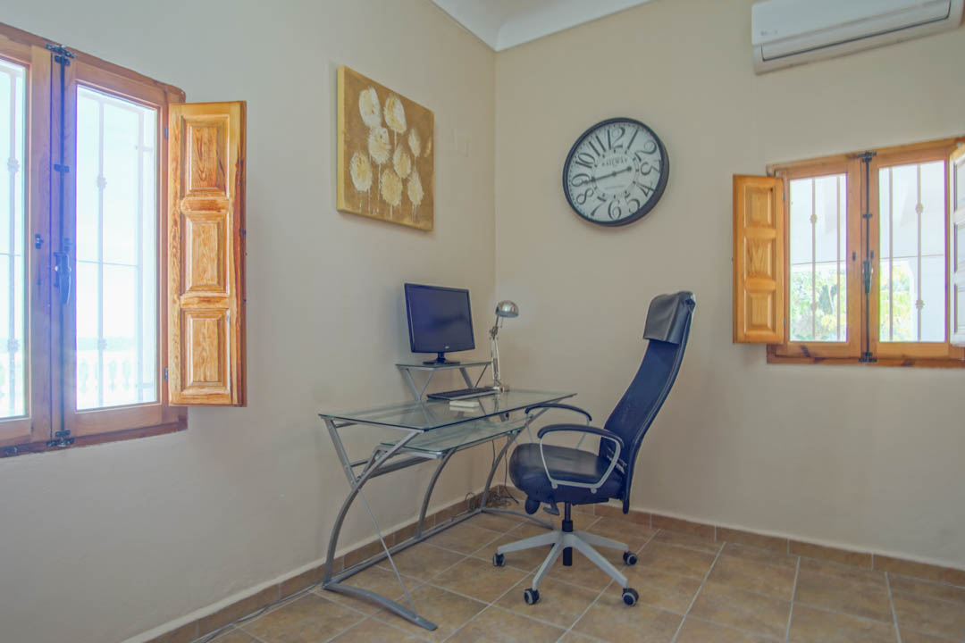 Espacio de oficina funcional con escritorio de cristal, ventanas de madera con contraventanas tradicionales y suelos de gres rústico.