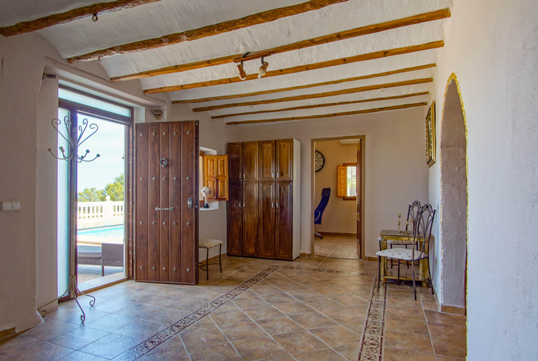 Hall de entrada rústico con vigas de madera a la vista, suelos cerámicos decorados y una puerta de madera maciza que conecta directamente con la zona de piscina exterior.