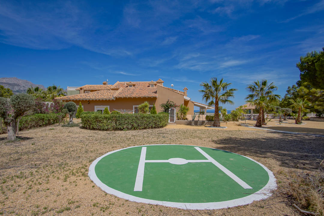 Helipuerto privado ubicado en la parte trasera de la propiedad, rodeado de jardines mediterráneos con palmeras y vistas a la arquitectura tradicional de la villa.