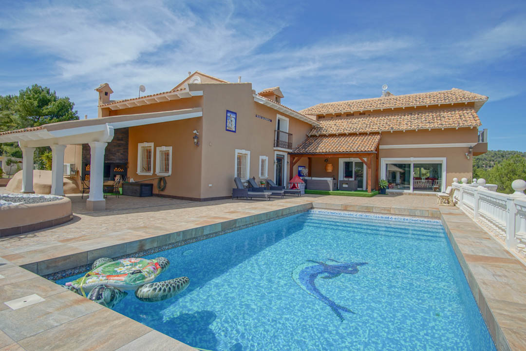 Piscina con mosaico decorativo rodeada de una amplia terraza de piedra, zona de barbacoa bajo porche con columnas blancas y arquitectura tradicional mediterránea.