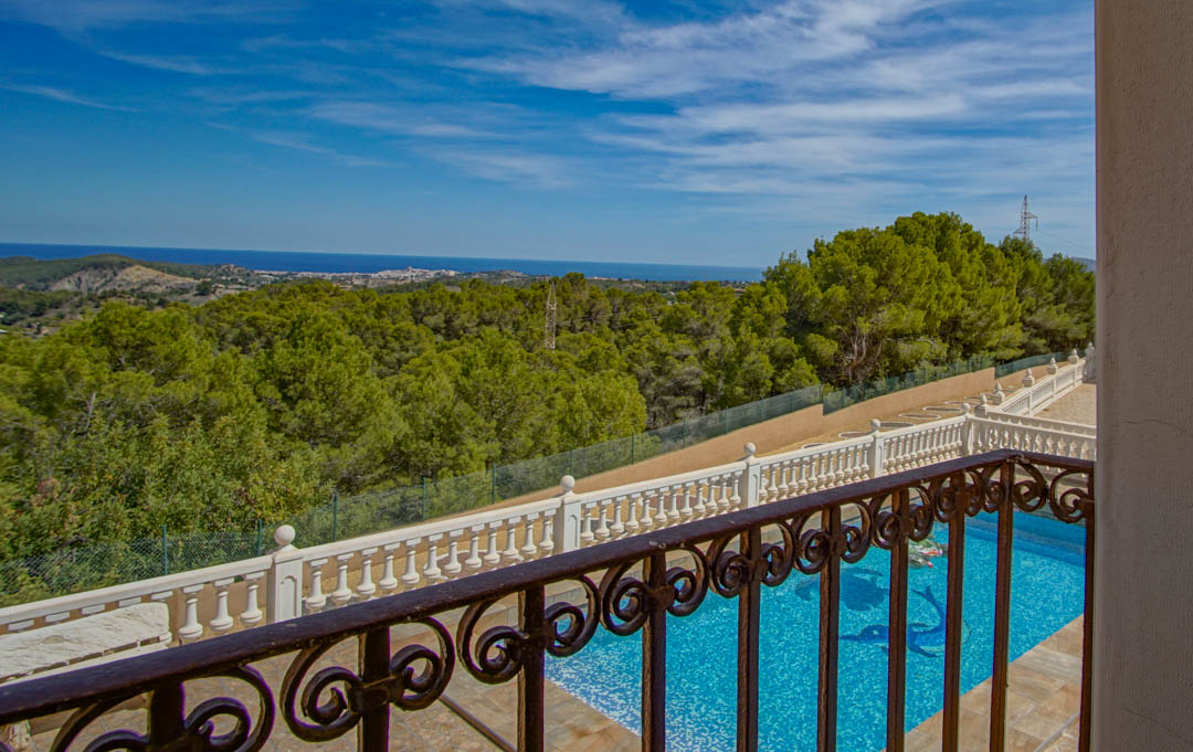 Vista desde el balcón con barandilla de hierro forjado hacia la piscina con mosaico, rodeada de pinos y vistas panorámicas al mar Mediterráneo.