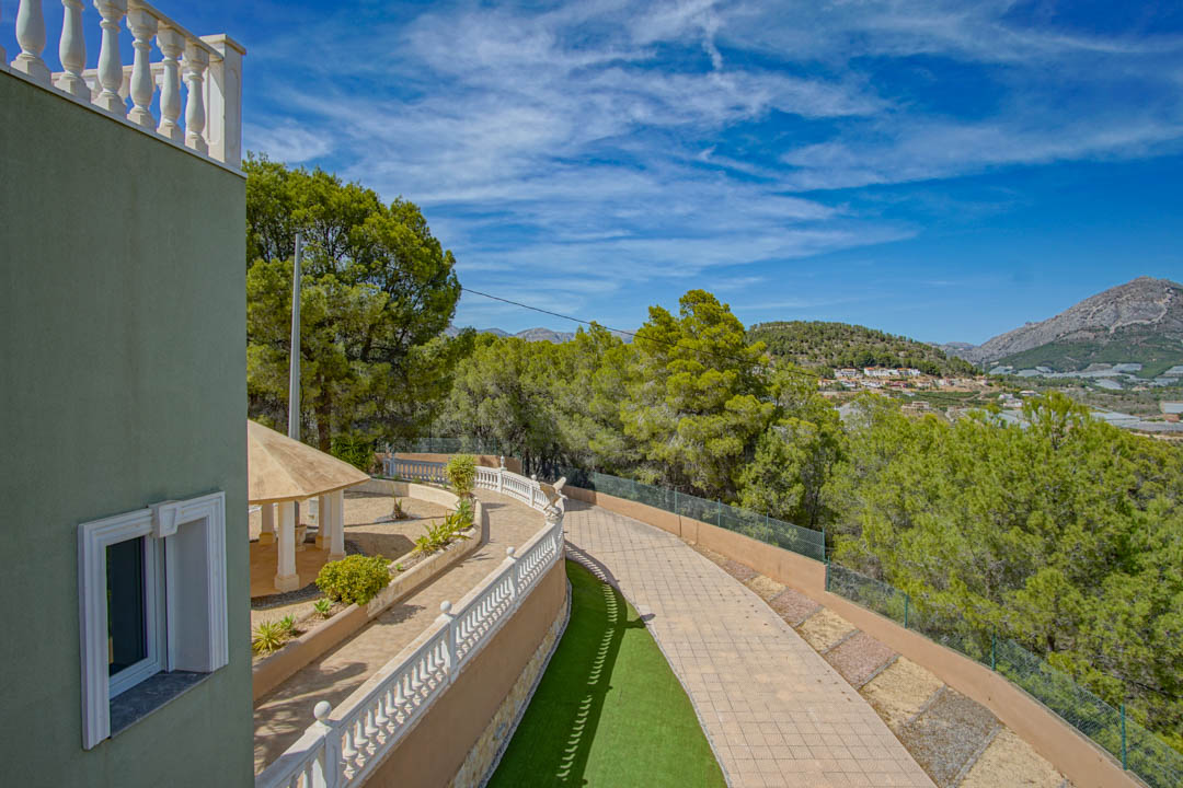 Vista exterior del camino pavimentado y balaustradas blancas. Incluye un pabellón techado junto a pinos maduros y amplias vistas a las montañas de La Nucía.